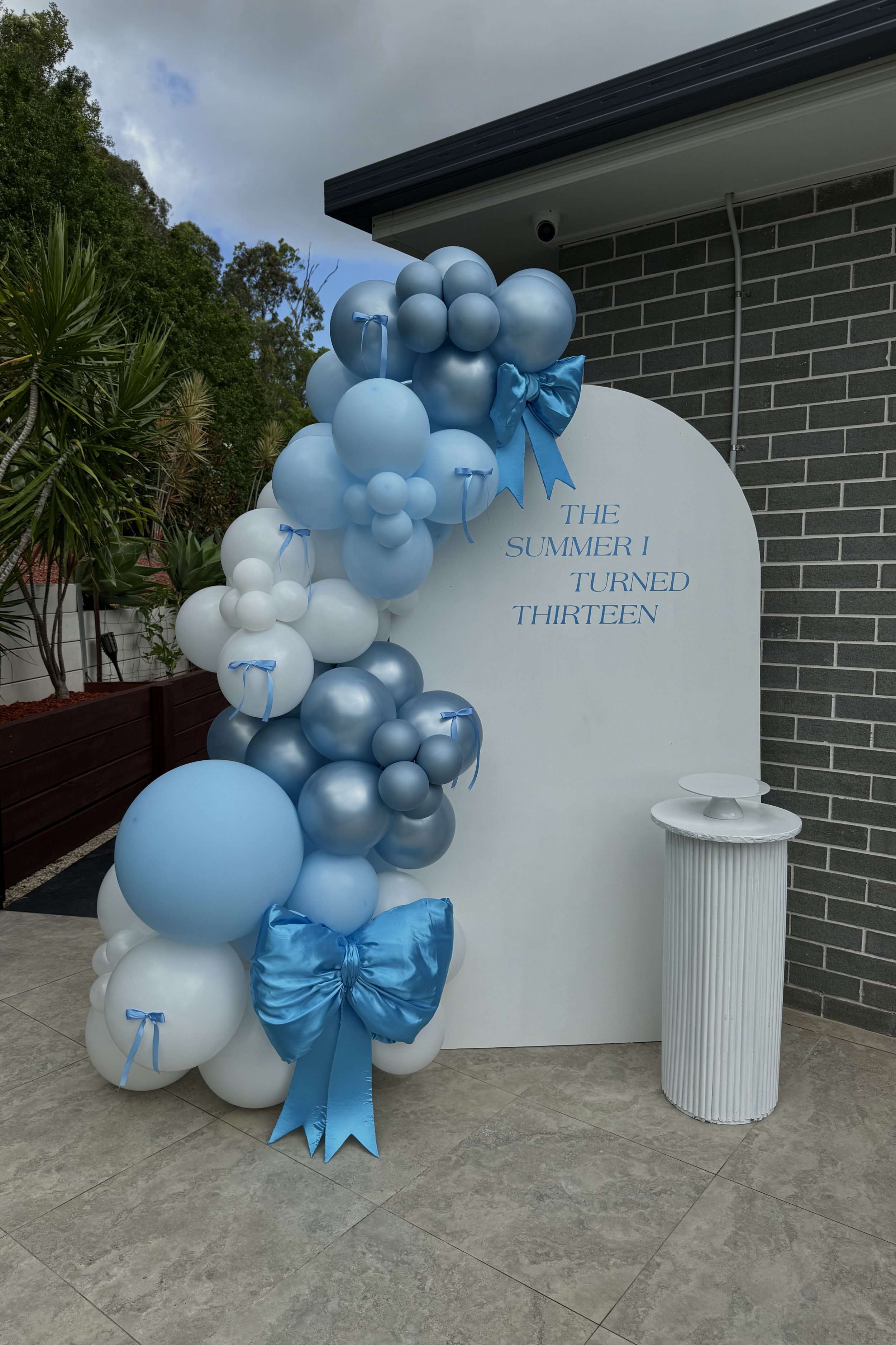 Blue and white balloon arrangement with bows and a large bow at the bottom, set against a white backdrop with the quote 'The summer I turned thirteen' in blue lettering.