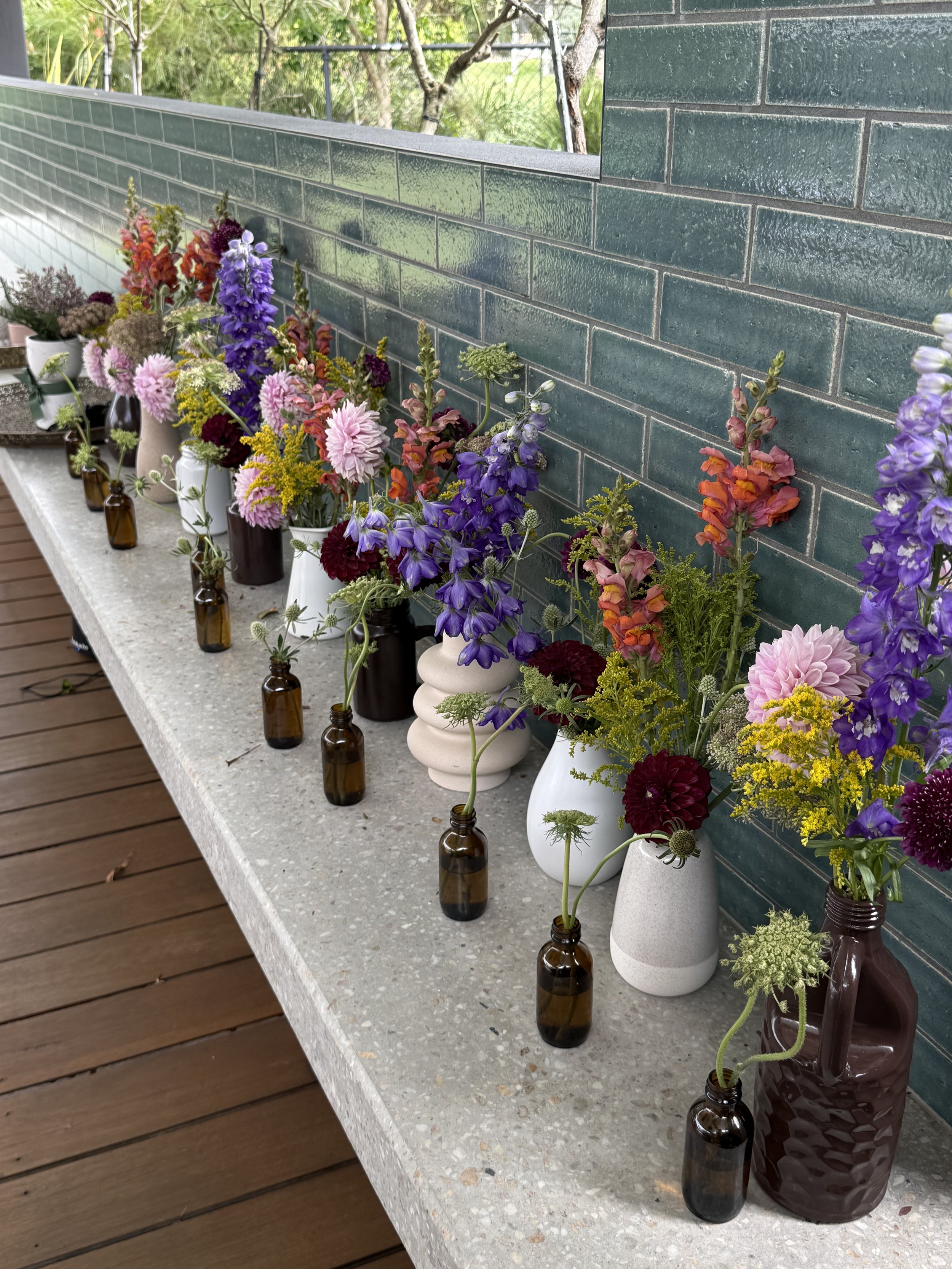 A variety of colorful flowers arranged in vases and small bottles. The flowers include pink dahlias, purple delphiniums, red and orange snapdragons, and yellow flowers, with greenery mixed in.