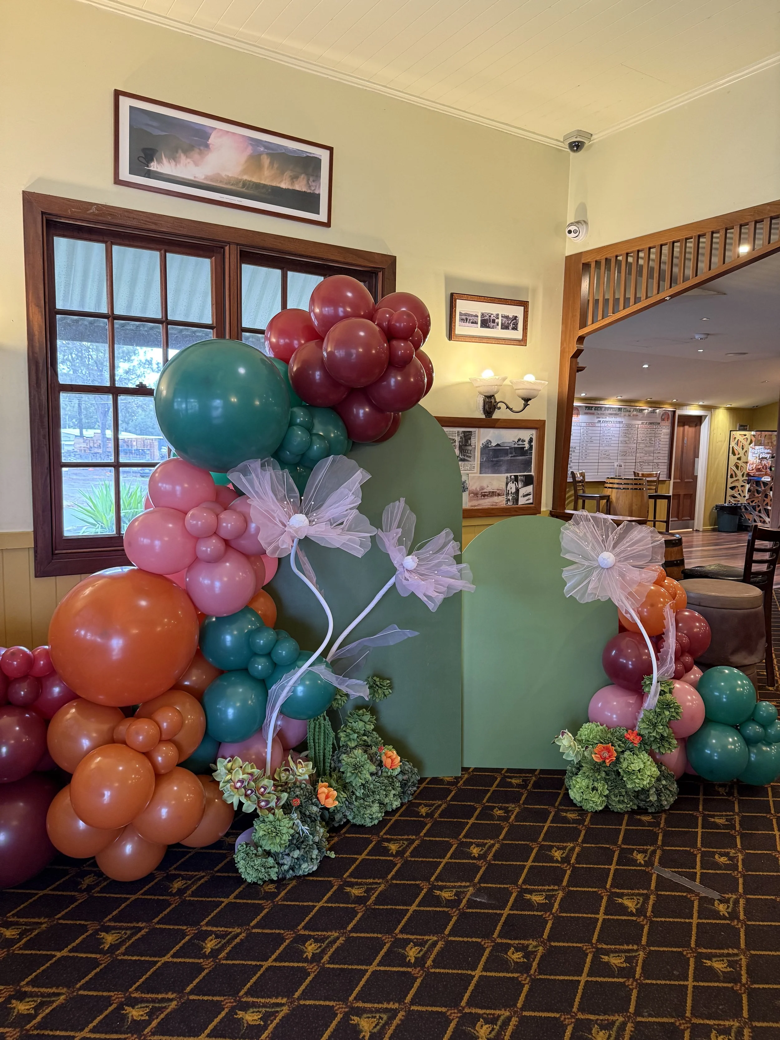 Melbourne cup themed set-up with burnt orange, dusty pink, sage green and wine colour balloon garland surrounded by giant tule flowers, floral arrangements with pops or orange on a double arch display giving a floral autumn feel.