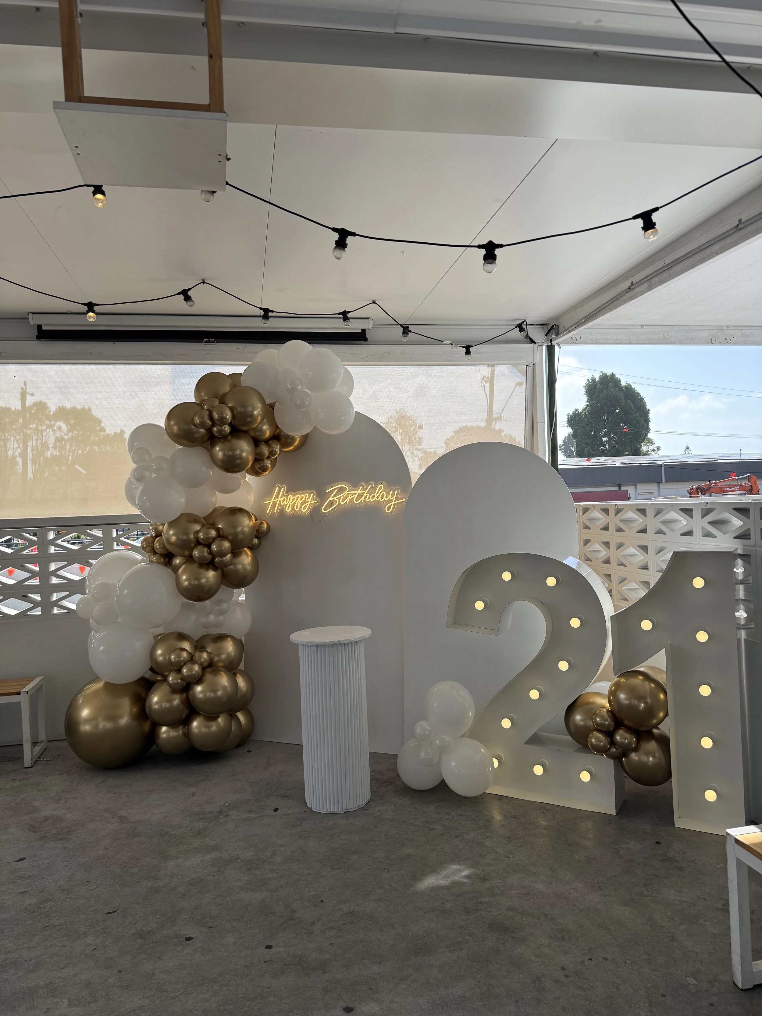 Decorated birthday celebration setup with large illuminated number 21, white and gold balloons, a white pedestal, and a neon sign that says 'Happy Birthday'.