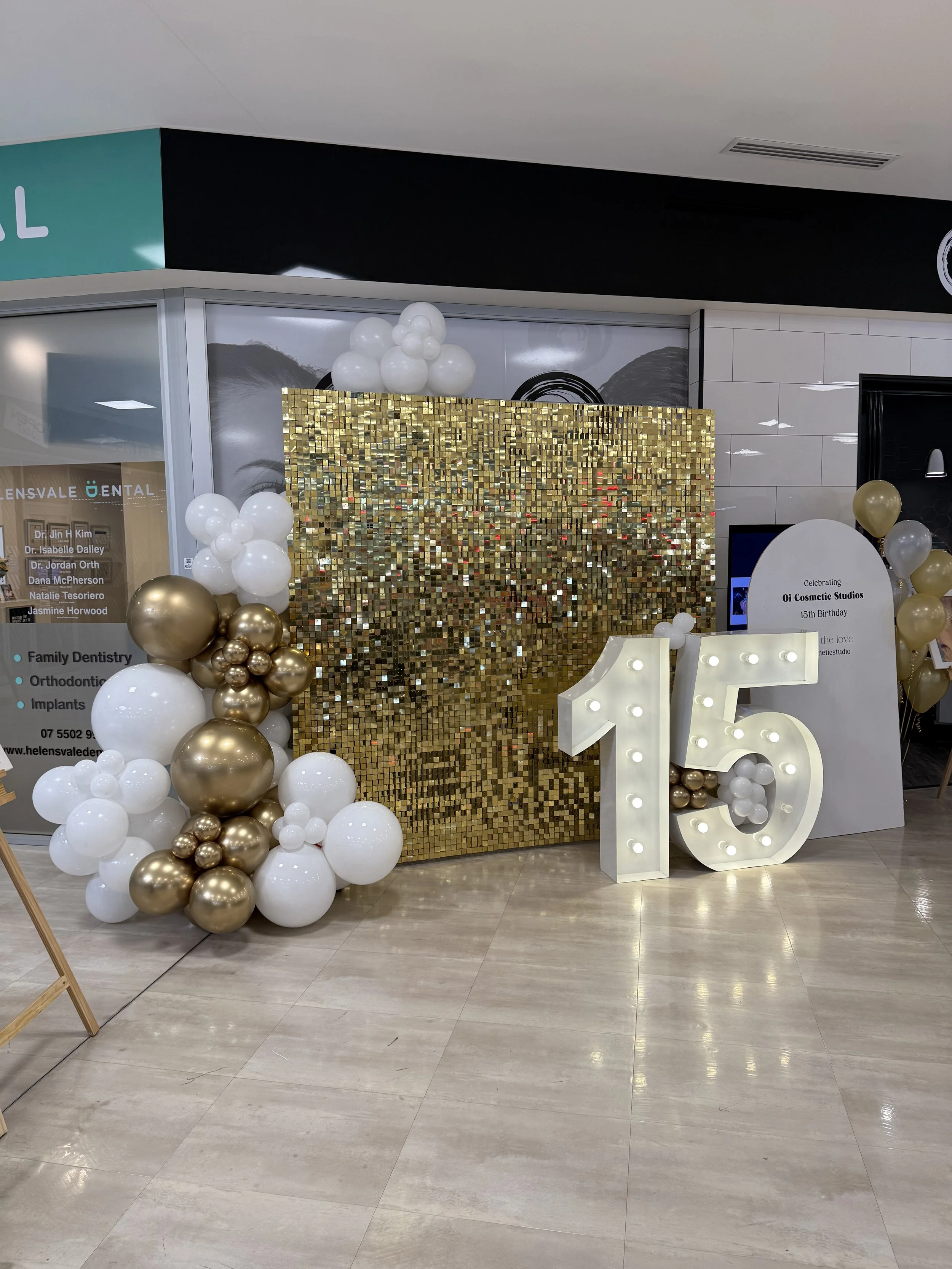 Decorative setup celebrating the 15th birthday of a Cosmetic Studios, featuring a large gold shimmer wall backdrop, a white and gold ballon garland and a large light up number 15.