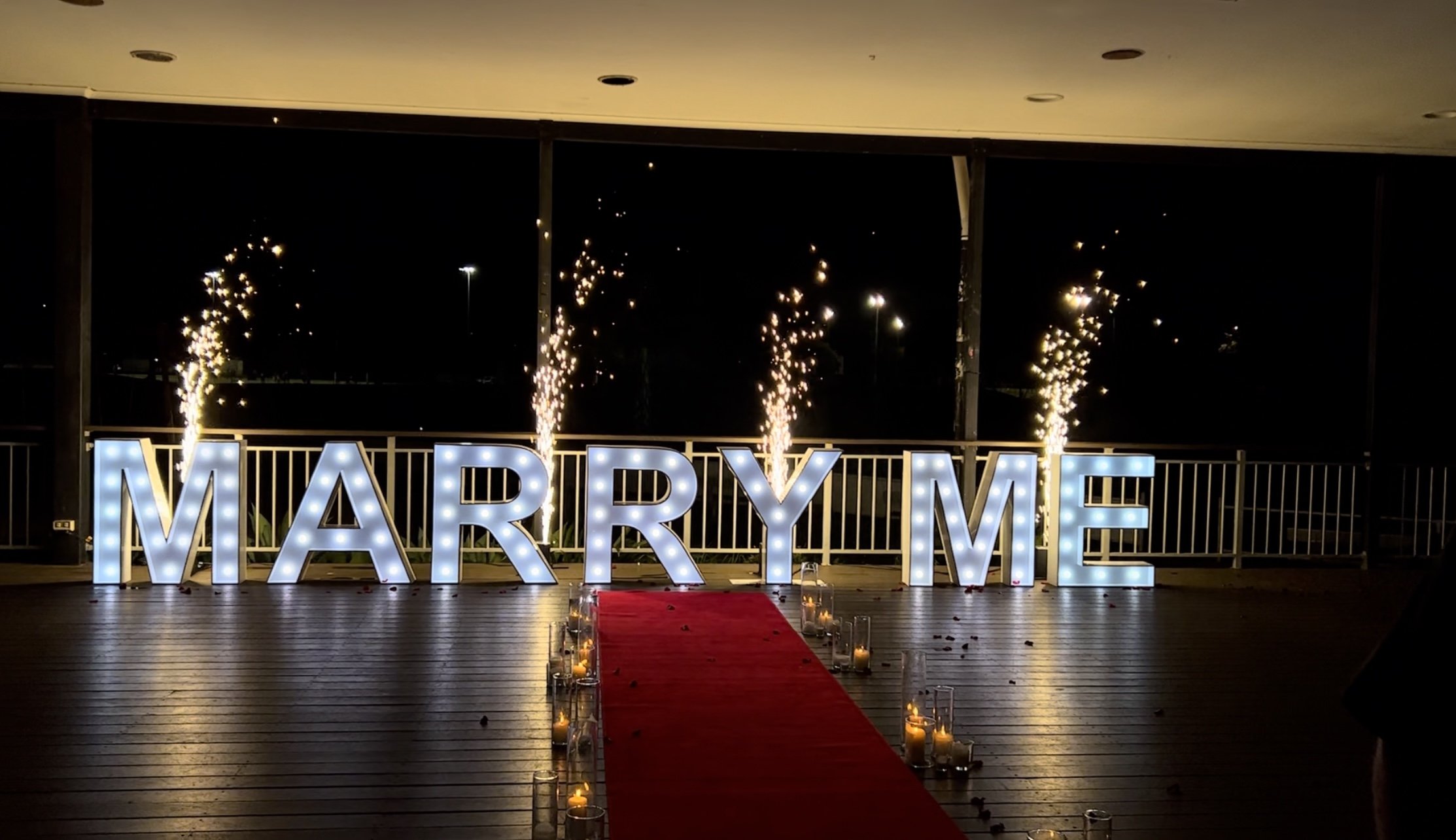 Large light up letters spelling 'MARRY ME' on a deck with sparklers and candles, a red carpet for a proposal.