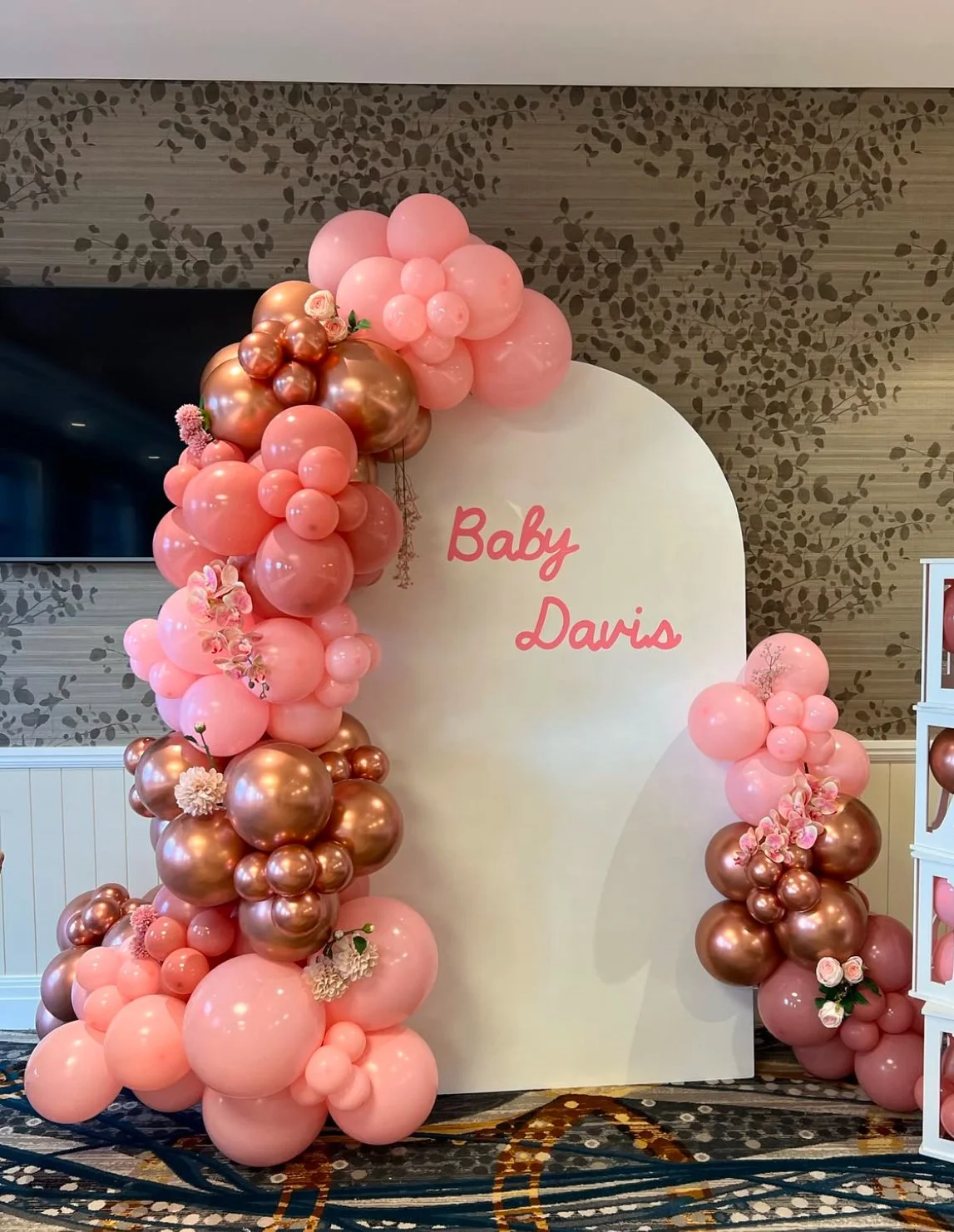 Decorative baby shower backdrop with pink and rose gold balloons and flowers, with the words "Baby Davis" written on it.