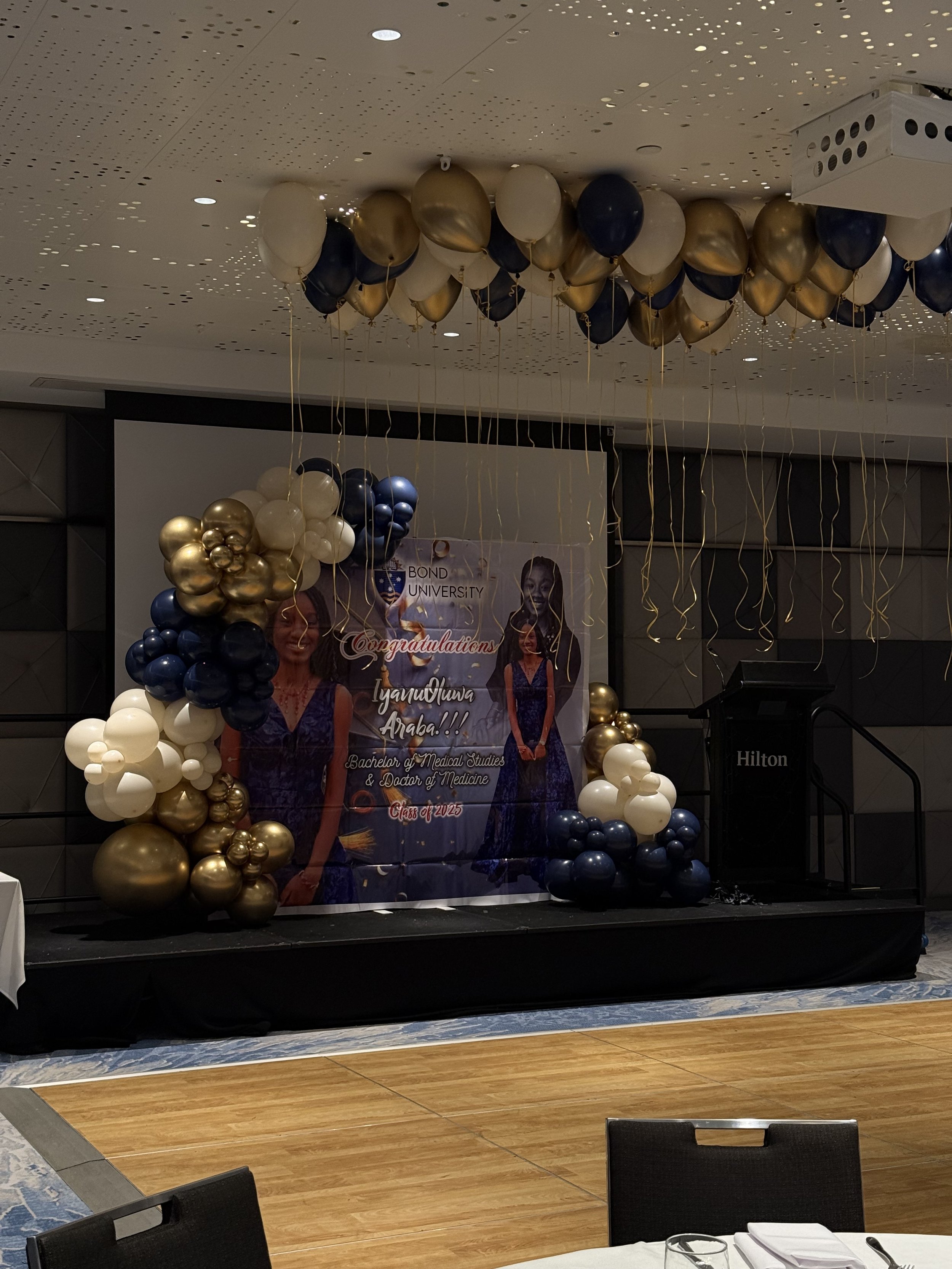 Balloon arch with gold, navy, and white balloons, decorated stage with a banner for a graduation celebration, and a podium at the Hilton hotel.
