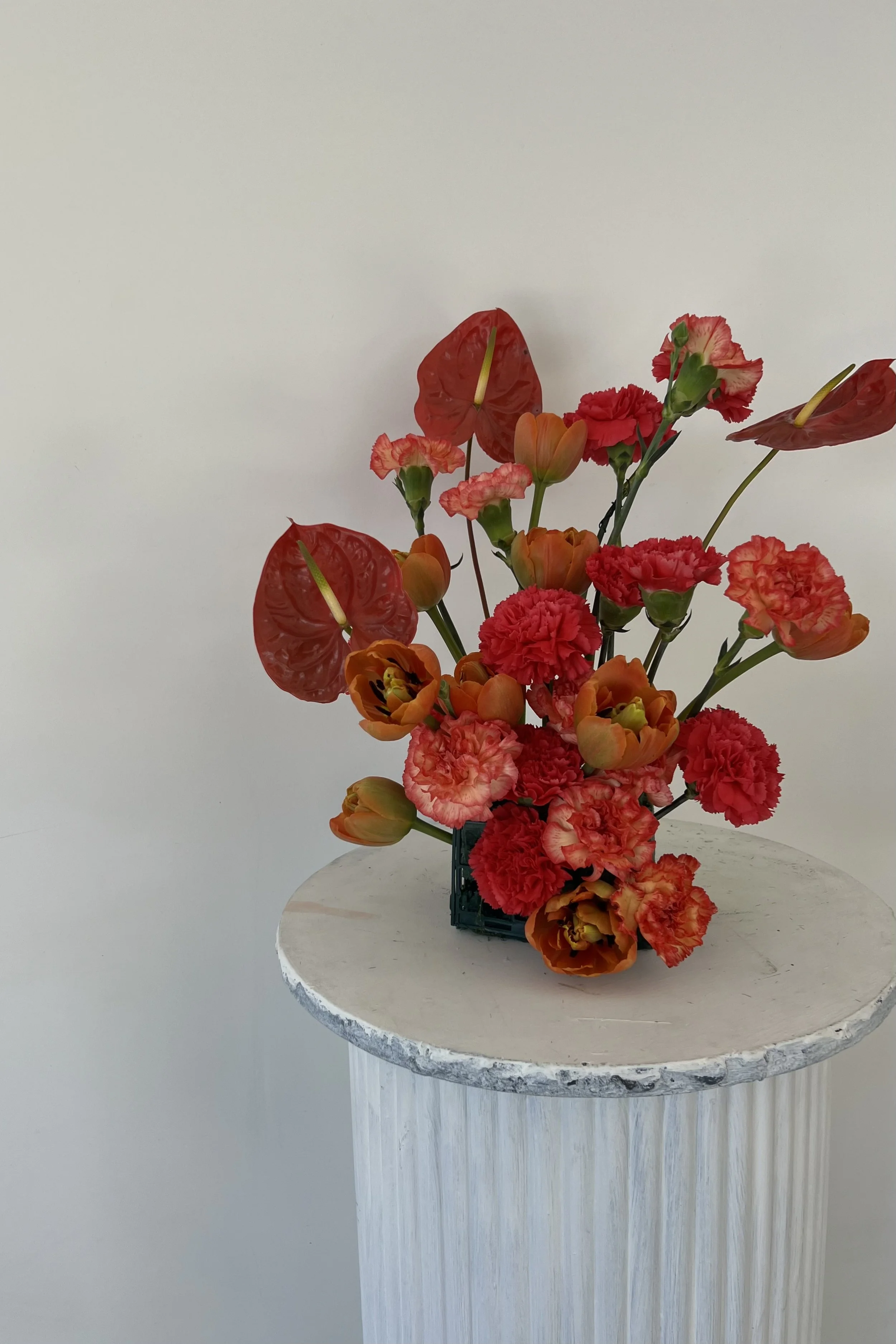 A floral arrangement with red anthuriums, orange tulips, and red carnations in a small black vase on a white pedestal against a plain background.
