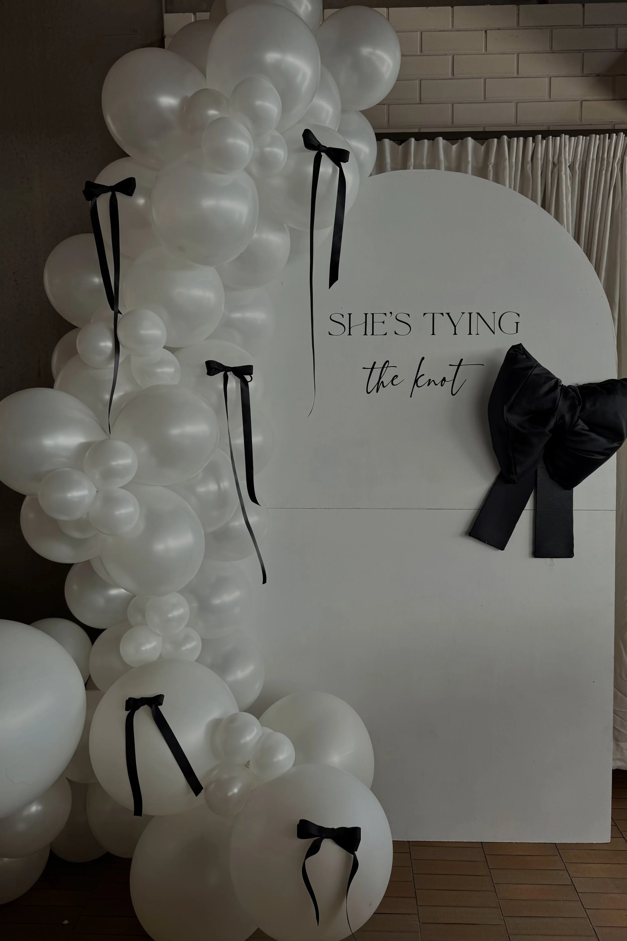 A white circular backdrop decorated with black bows and balloons, with the words "SHE'S TYING the knot" written on it, indicating a wedding or bridal celebration.