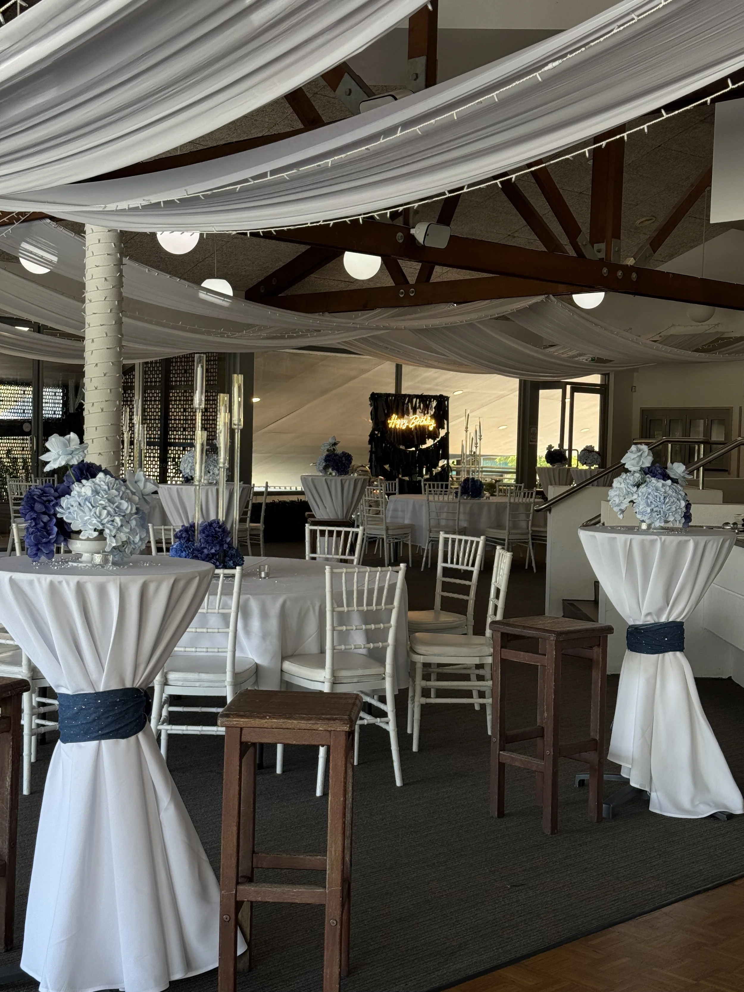 Indoor event space decorated with white drapes, floral centerpieces, and white chairs with blue sashes. There are round tables, some with floral arrangements, and a background featuring a stage with a neon sign that says 'Happy Birthday'.