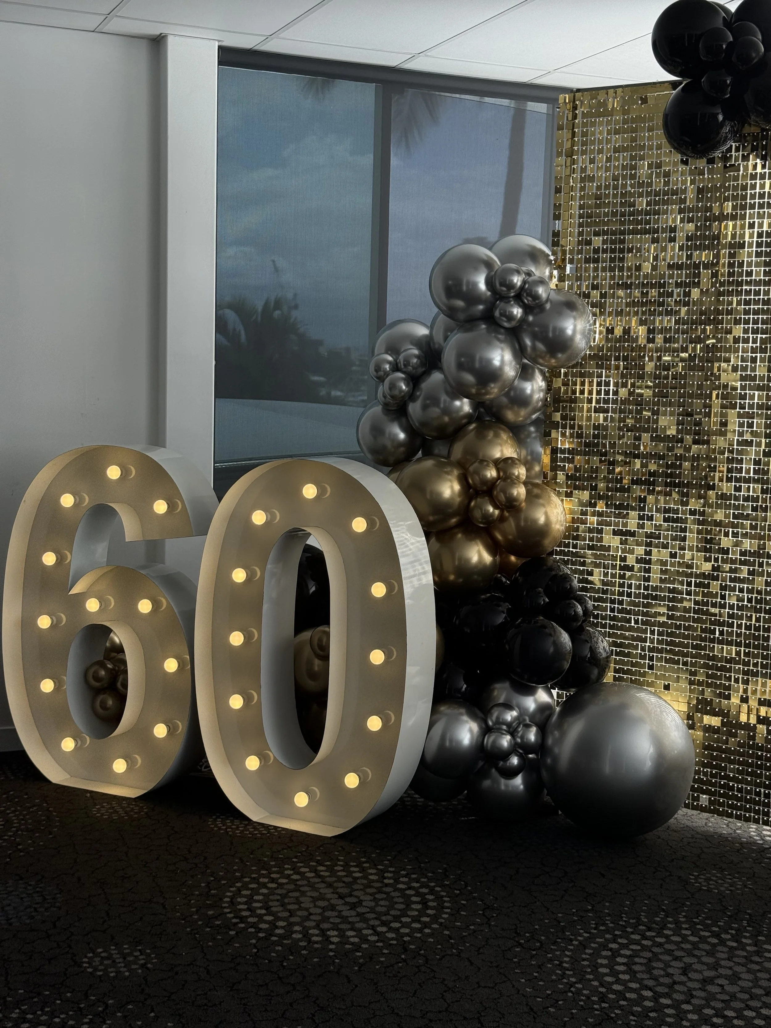 Decorative display with illuminated large numbers '60', arrangements of black, silver, and gold balloons, and a gold sequin backdrop, celebrating a 60th milestone.