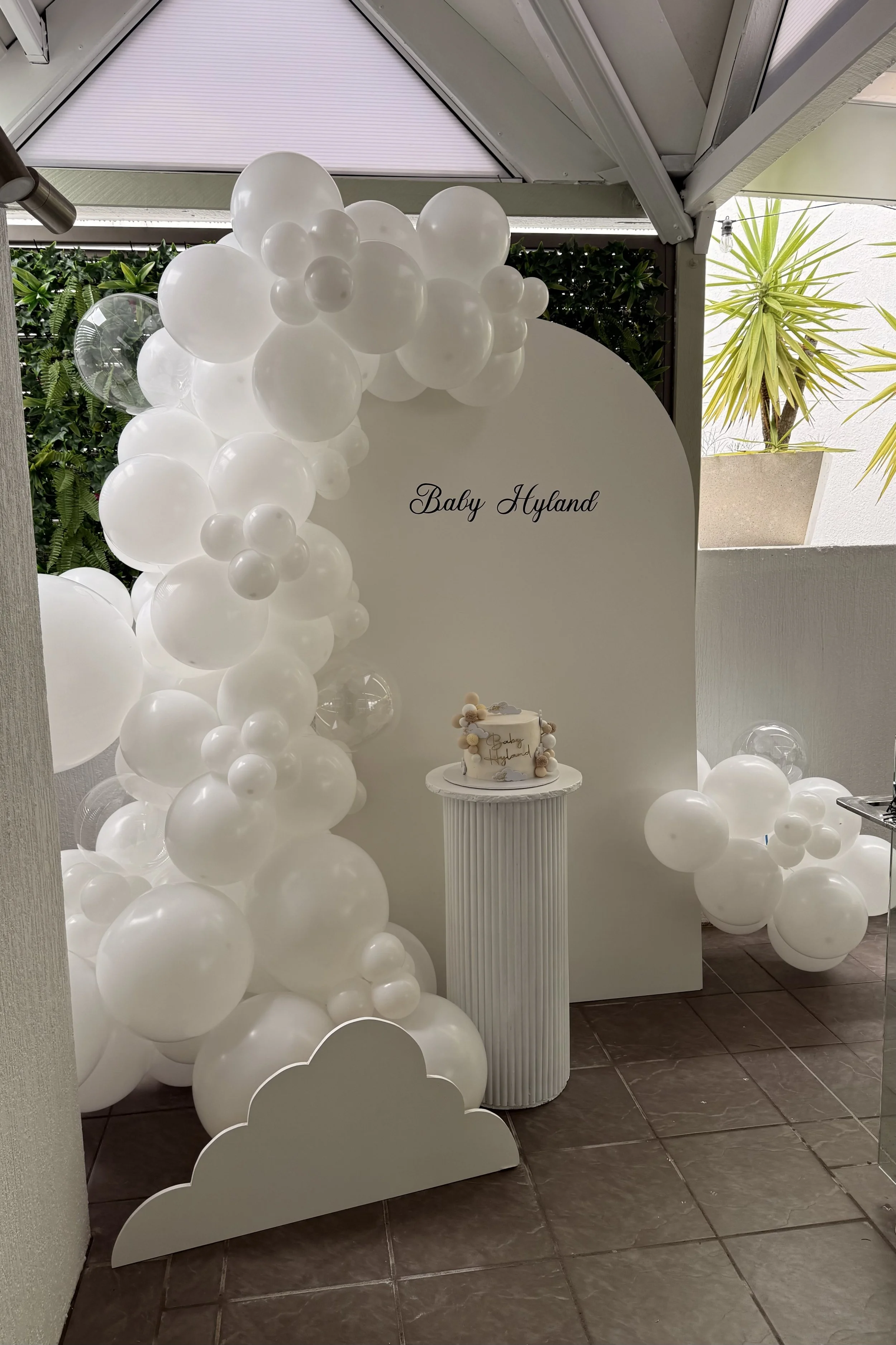 Baby shower decoration with white balloons, a white cake on a pedestal, and a white sign reading 'Baby Hyland' in cursive, set against a white backdrop