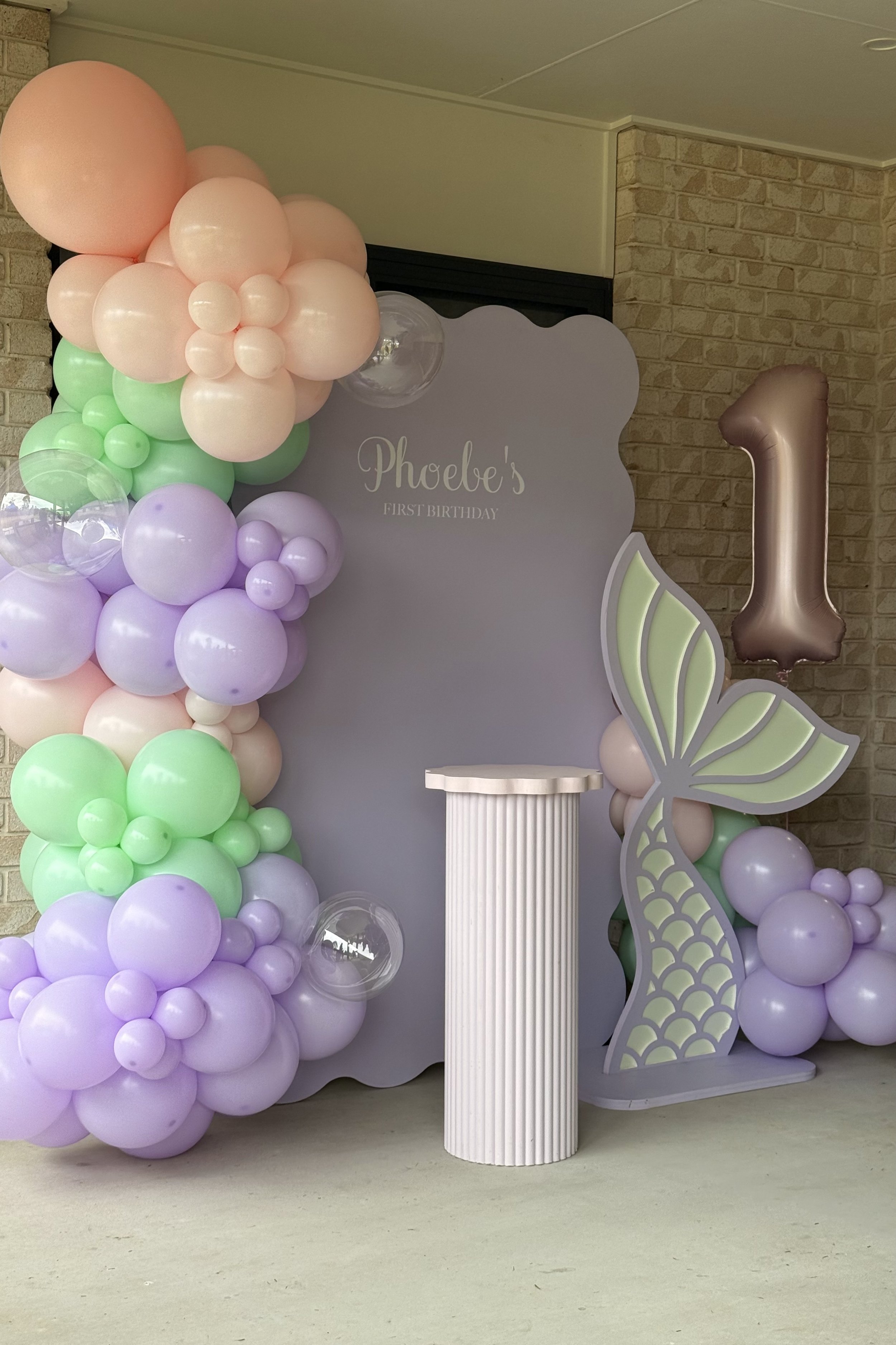 Decorations for a first birthday party with pastel balloons, a butterfly cutout, a large number 1 balloon, and a display board reading 'Phoebe's First Birthday.'