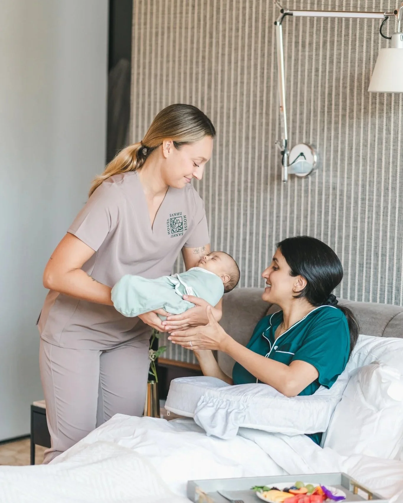 Sanhu House postpartum doula handing a newborn to a mother resting in bed