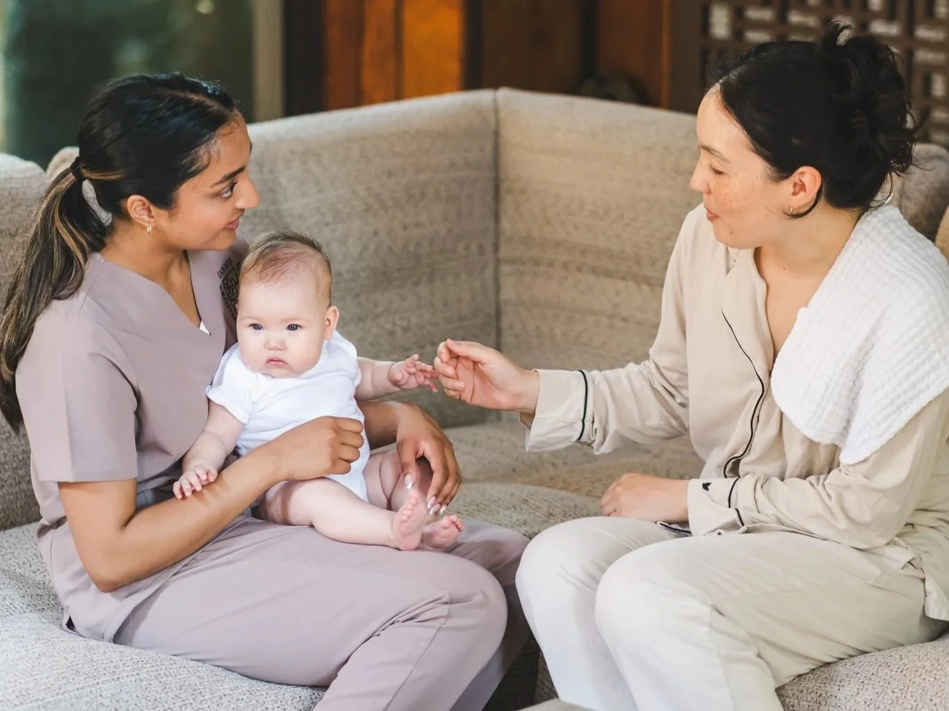 A woman in medical scrubs holding a baby while another woman in pajamas interacts with the baby on a couch in a living room.