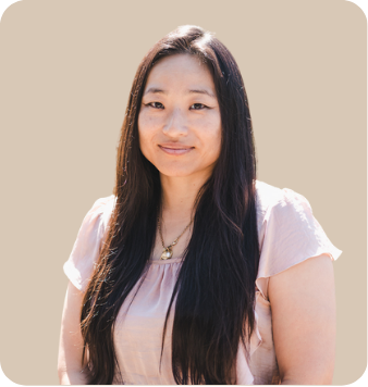 A woman with long dark hair wearing a light pink top and necklace, smiling at the camera against a plain background.