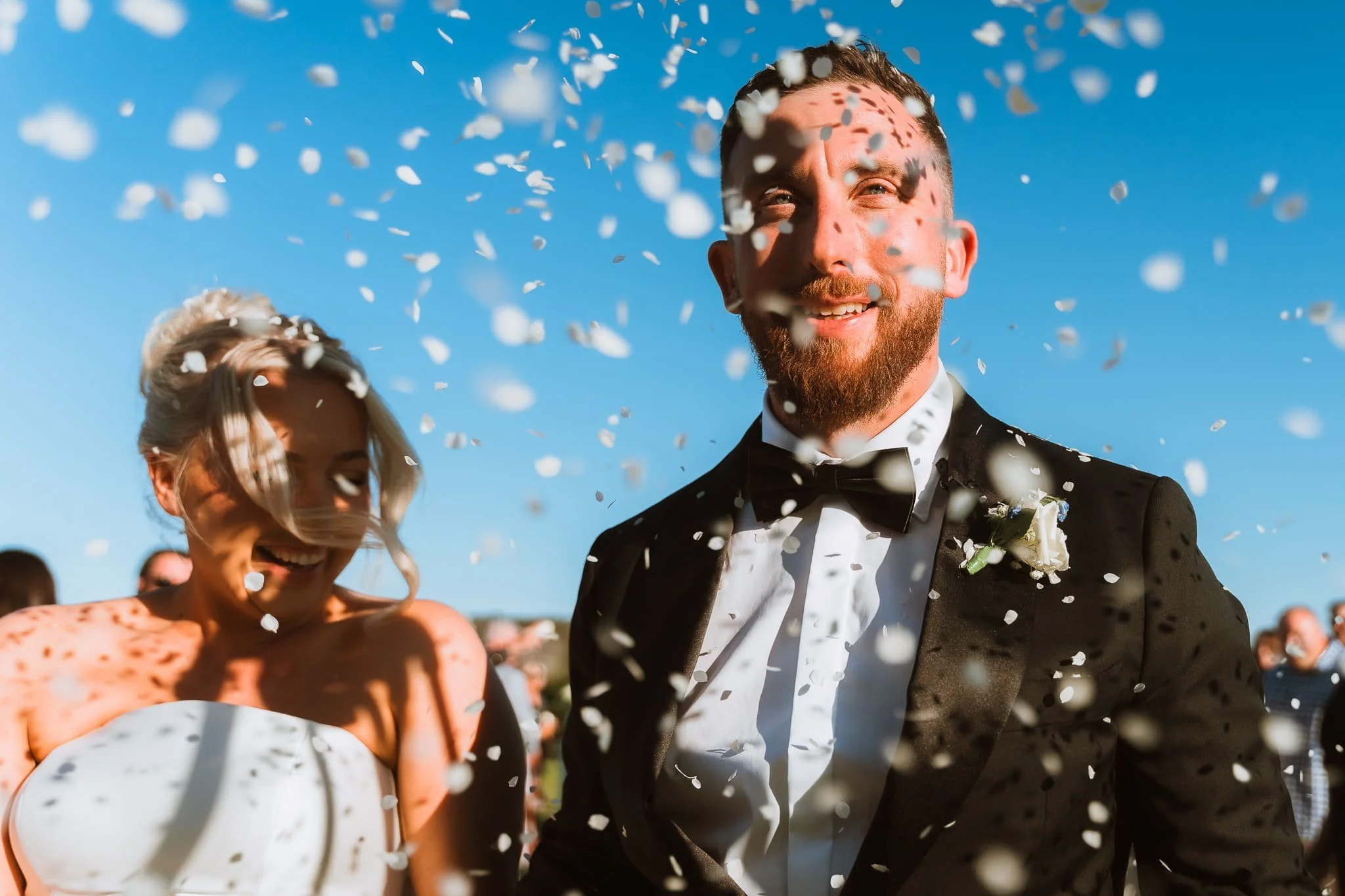 A man dressed in a tuxedo with a bow tie, likely at a wedding, is smiling among falling confetti during an outdoor celebration on a clear sunny day.