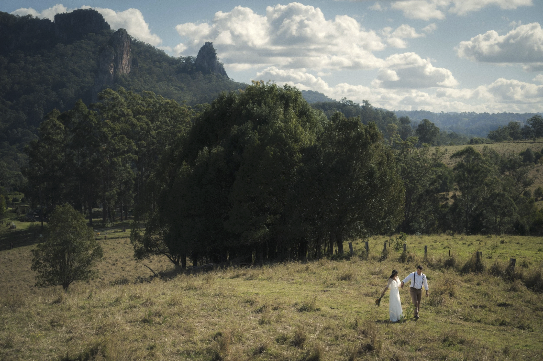 A couple walking hand in hand in an open grassy field, surrounded by trees and distant mountains under a partly cloudy sky.