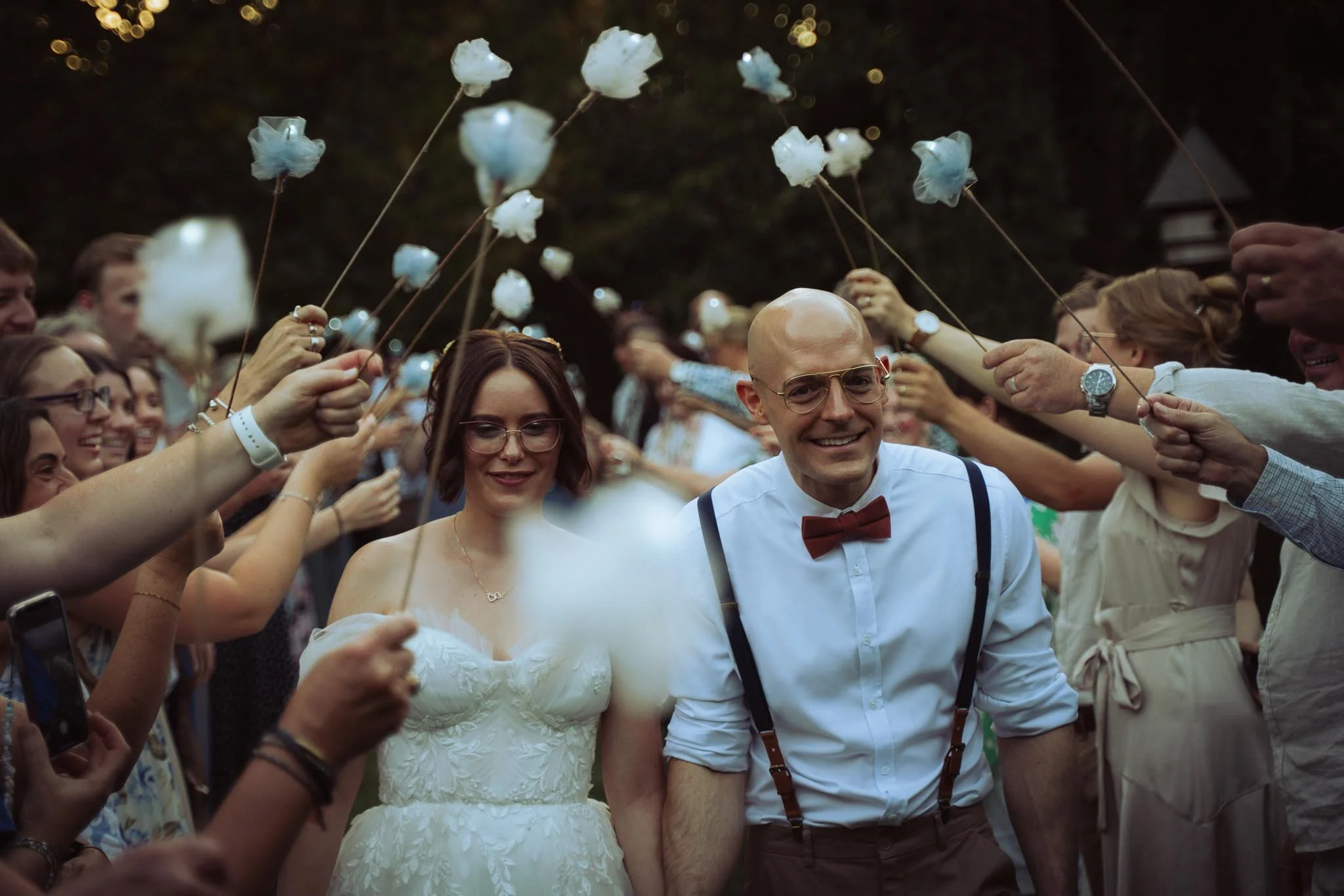 A couple, a woman in a white wedding dress and a man in a white shirt with suspenders and red bow tie, walking outdoors during a celebration. They are surrounded by people holding cotton candy on sticks, creating an arch for them to walk through. The couple appears happy, with the woman smiling softly and the man smiling widely.