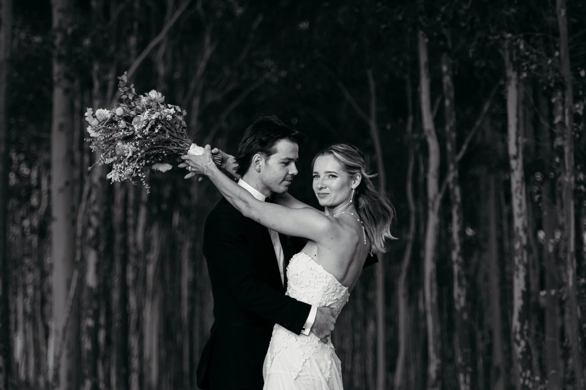 A black and white photo of a couple dancing outdoors, with trees in the background, the woman wearing a white lace dress and the man in a dark tuxedo, holding each other close.