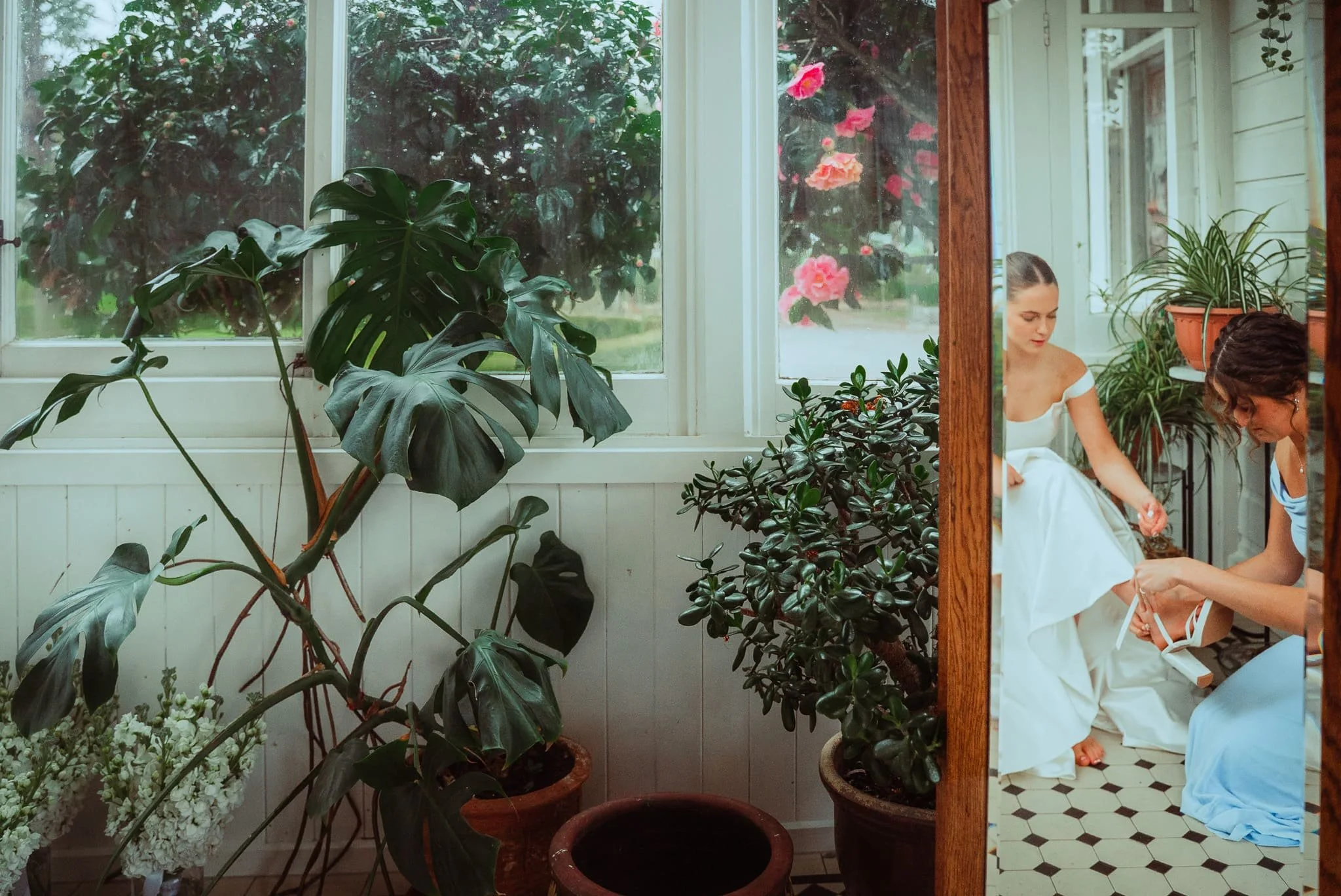 Two women preparing for a wedding indoors, surrounded by houseplants, with a large window showing outdoor greenery and pink flowers.