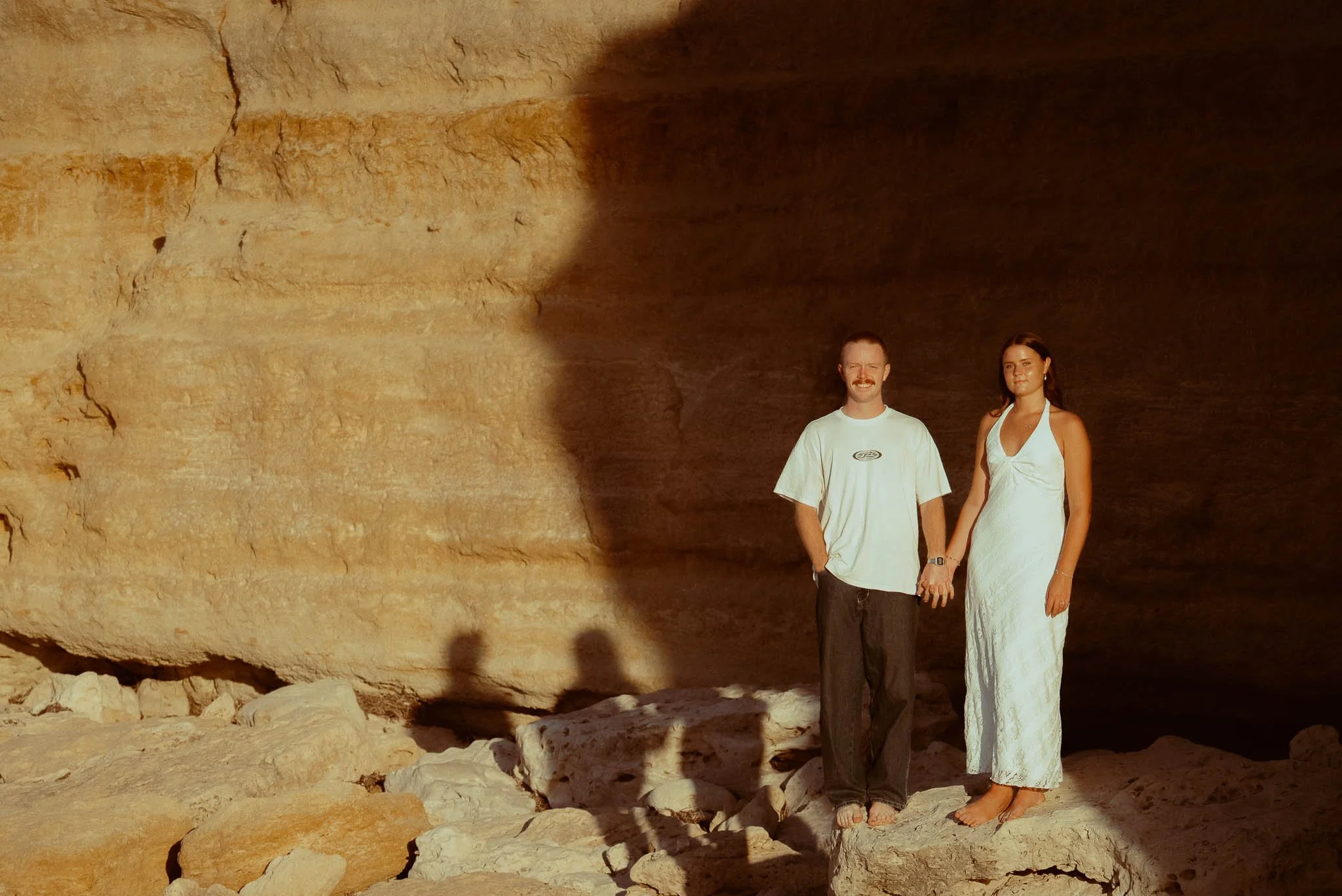 A man and woman standing inside a large cave with rocky walls and ground, sunlight casting shadows on the scene.