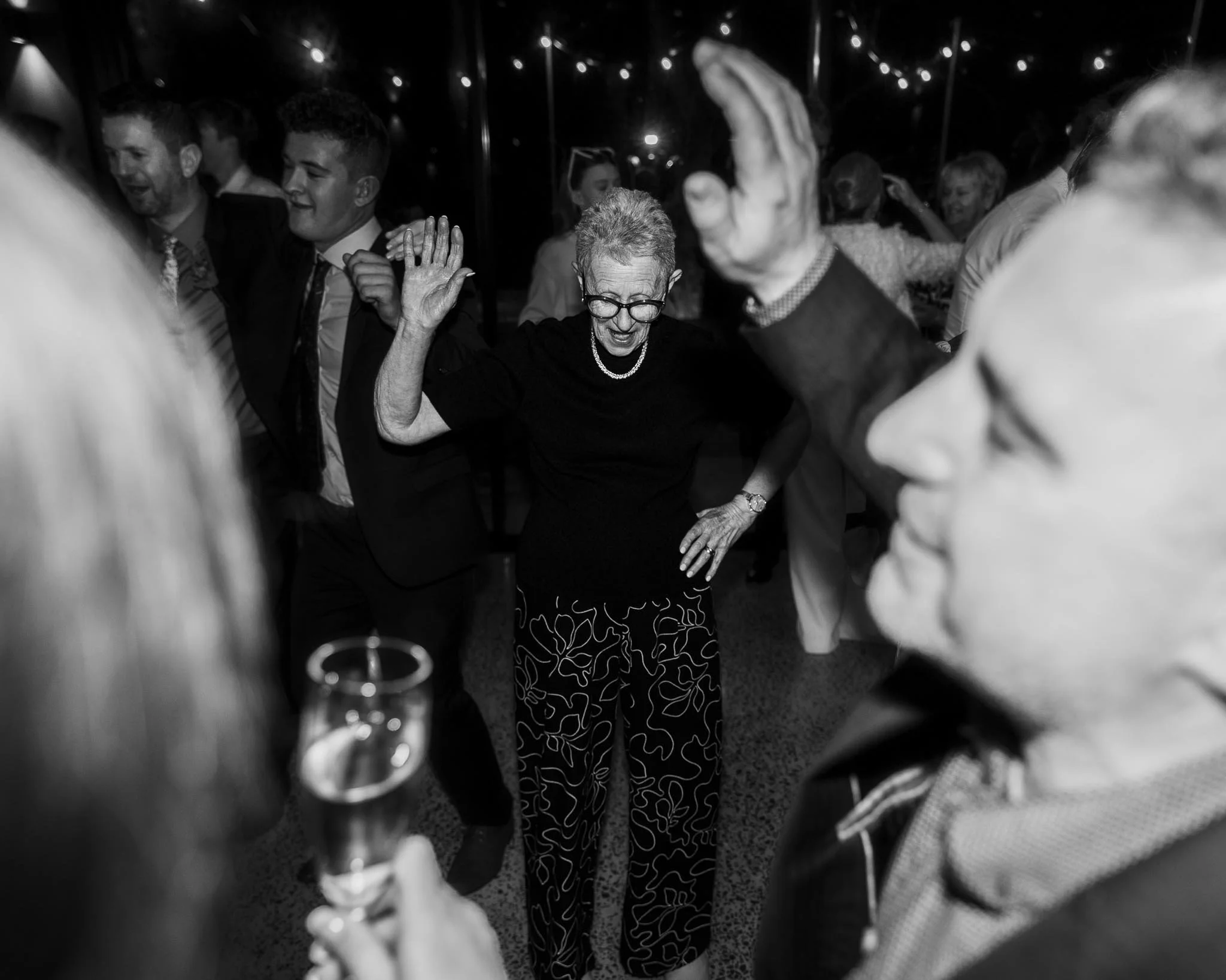 People dancing at a lively party or celebration, including an elderly woman in the center, wearing glasses, a black top, and patterned pants, surrounded by smiling guests.