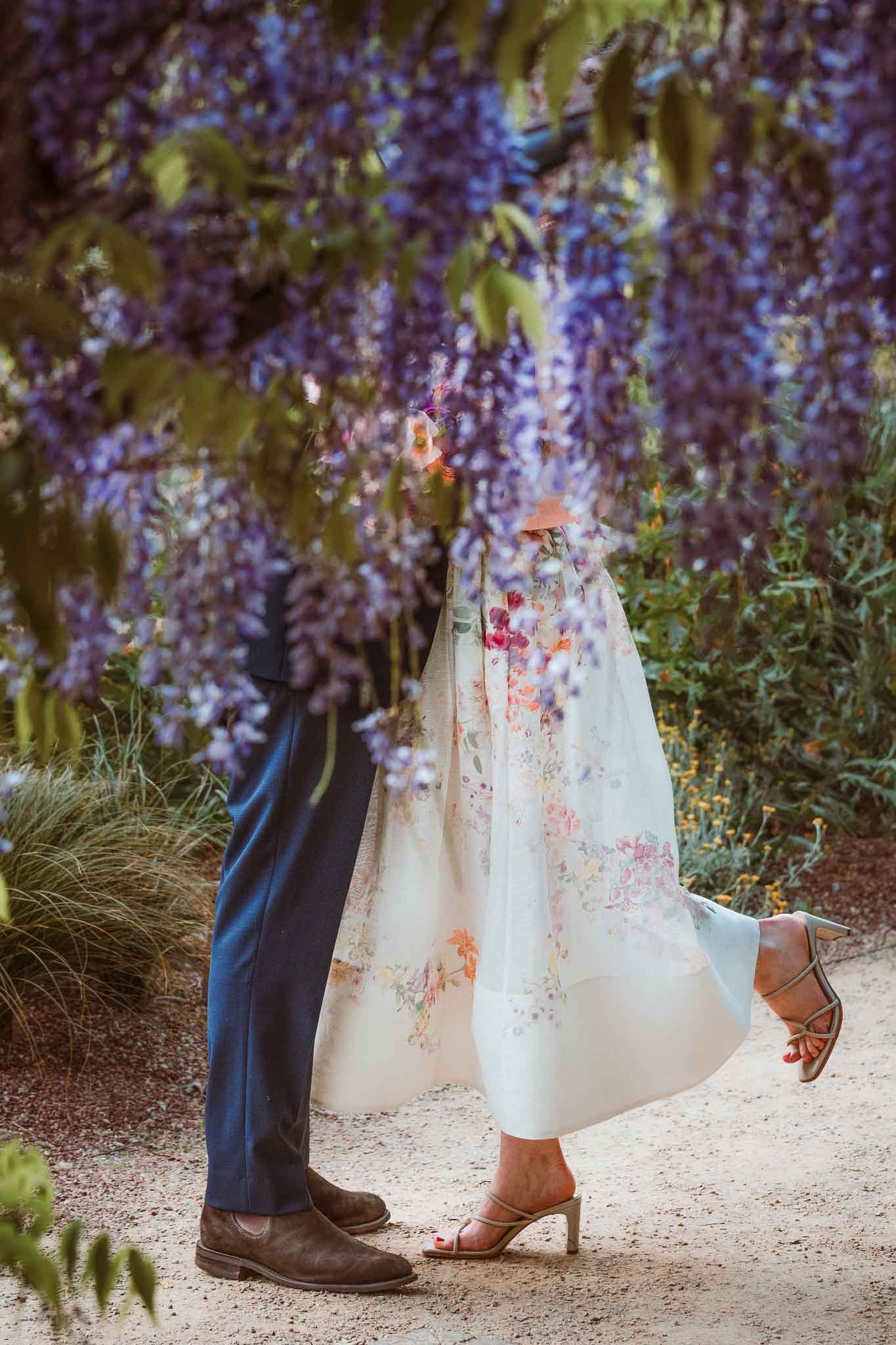 Close-up of a couple's lower bodies, with the woman wearing a long floral dress and high heels, and the man wearing dark pants and brown boots, standing on a dirt path surrounded by green foliage and purple flowering plants.