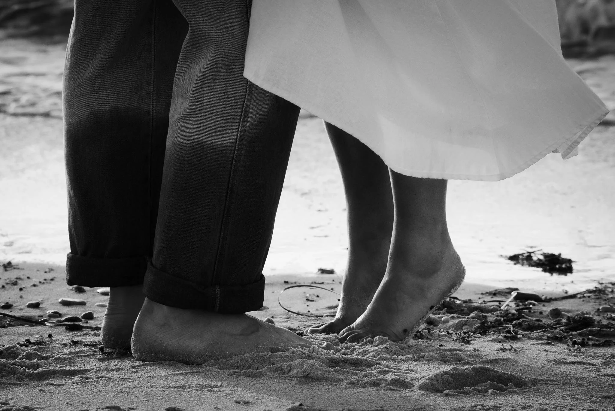 Close-up of a couple's feet on the beach, with one person barefoot and the other in rolled-up jeans, and a woman in a white dress.