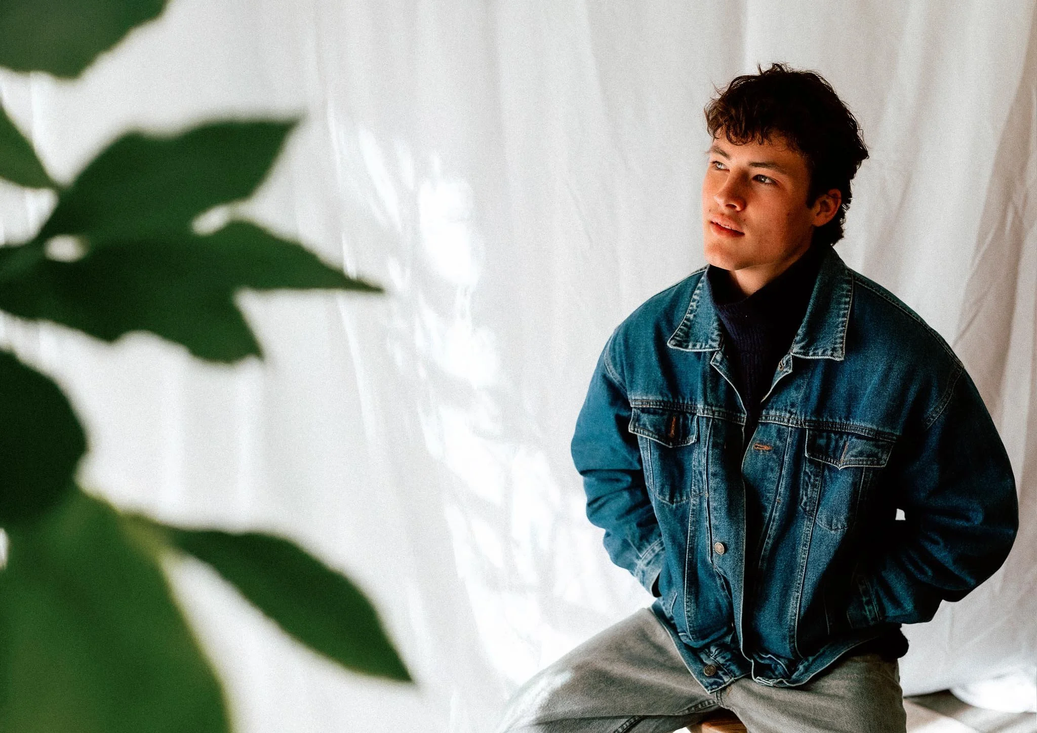 A young man with dark wavy hair, wearing a denim jacket and gray pants, sitting on a stool against a white fabric backdrop, with green leaves partially visible in the foreground.