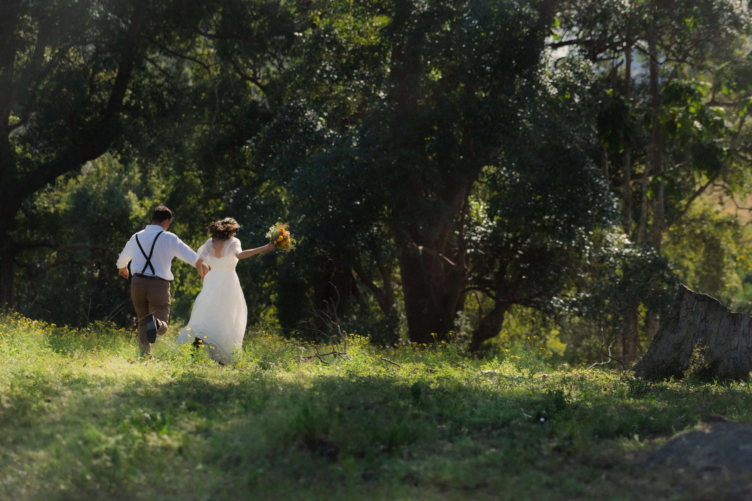 A bride in a white wedding dress holding a bouquet walking in a grassy outdoor area, with a man in suspenders and brown pants. Trees with green foliage surround them, and they appear to be running or walking quickly.