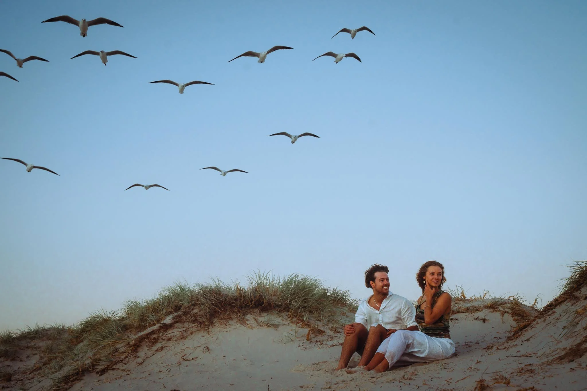 A couple sitting on sandy dunes with grass, looking happy together, under a clear blue sky with seagulls flying overhead.