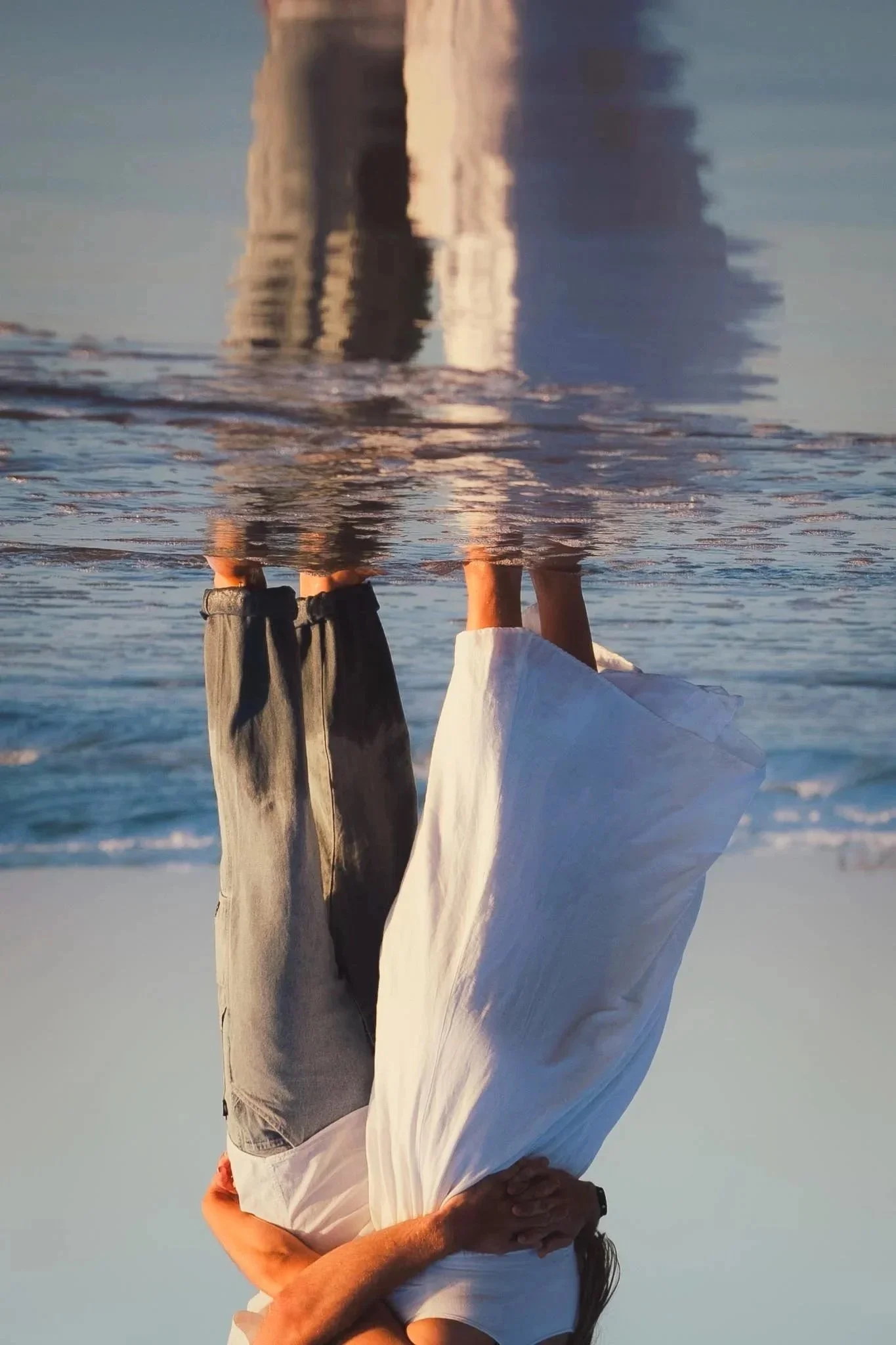 A person standing on the beach holding another person upside down in a handstand. The person being held is upside down with their head near the sand. The photo is taken at sunset or sunrise, reflecting the couple's image on the water.