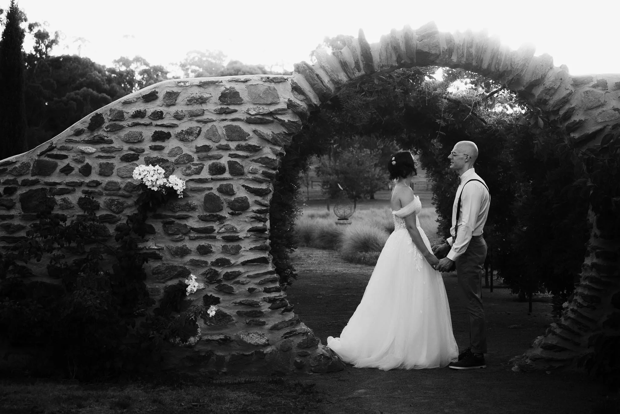 A black and white photograph of a bride and groom holding hands under a stone archway outdoors, with trees in the background.
