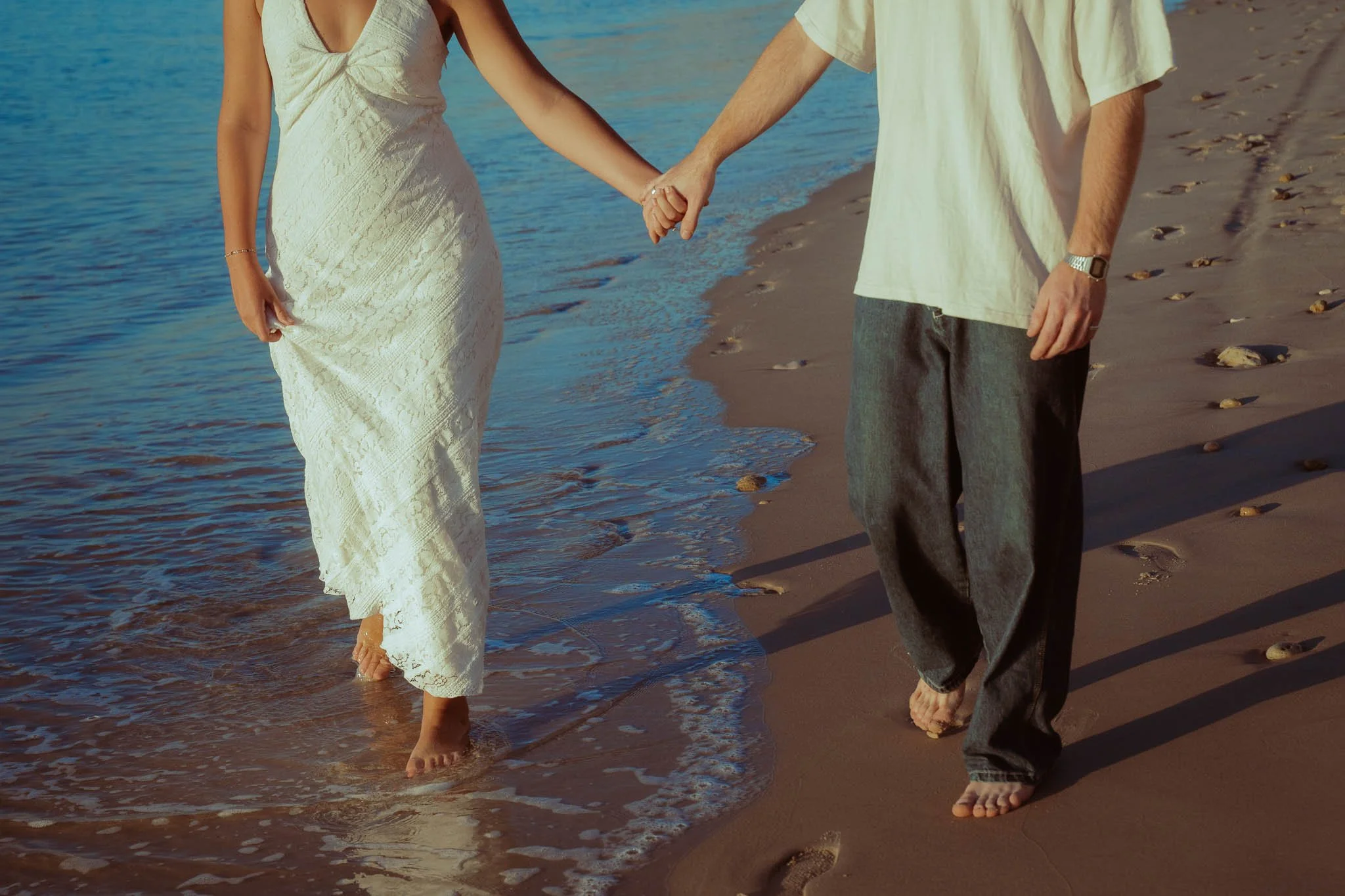 A couple holding hands while walking barefoot along the shoreline with gentle waves at sunset.