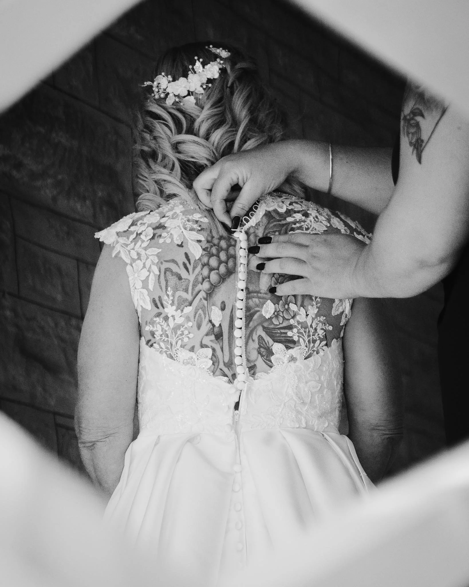 A woman in a wedding dress is buttoning the back of her gown while another person helps, with focus on their hands and the woman's intricate lace dress.