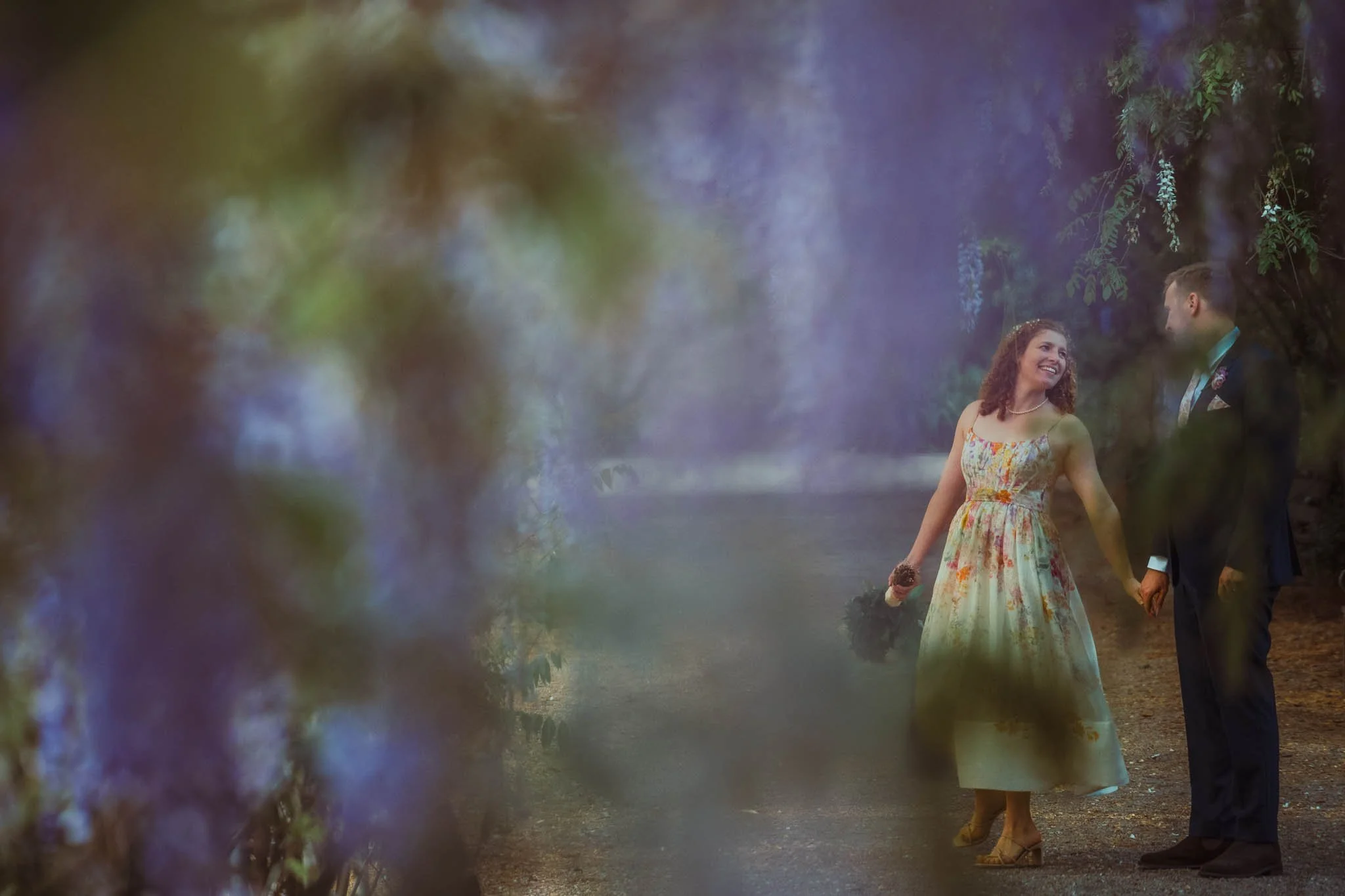 A bride and groom holding hands outdoors, with the bride wearing a colorful floral dress and the groom in a suit, surrounded by greenery and blurred foliage.