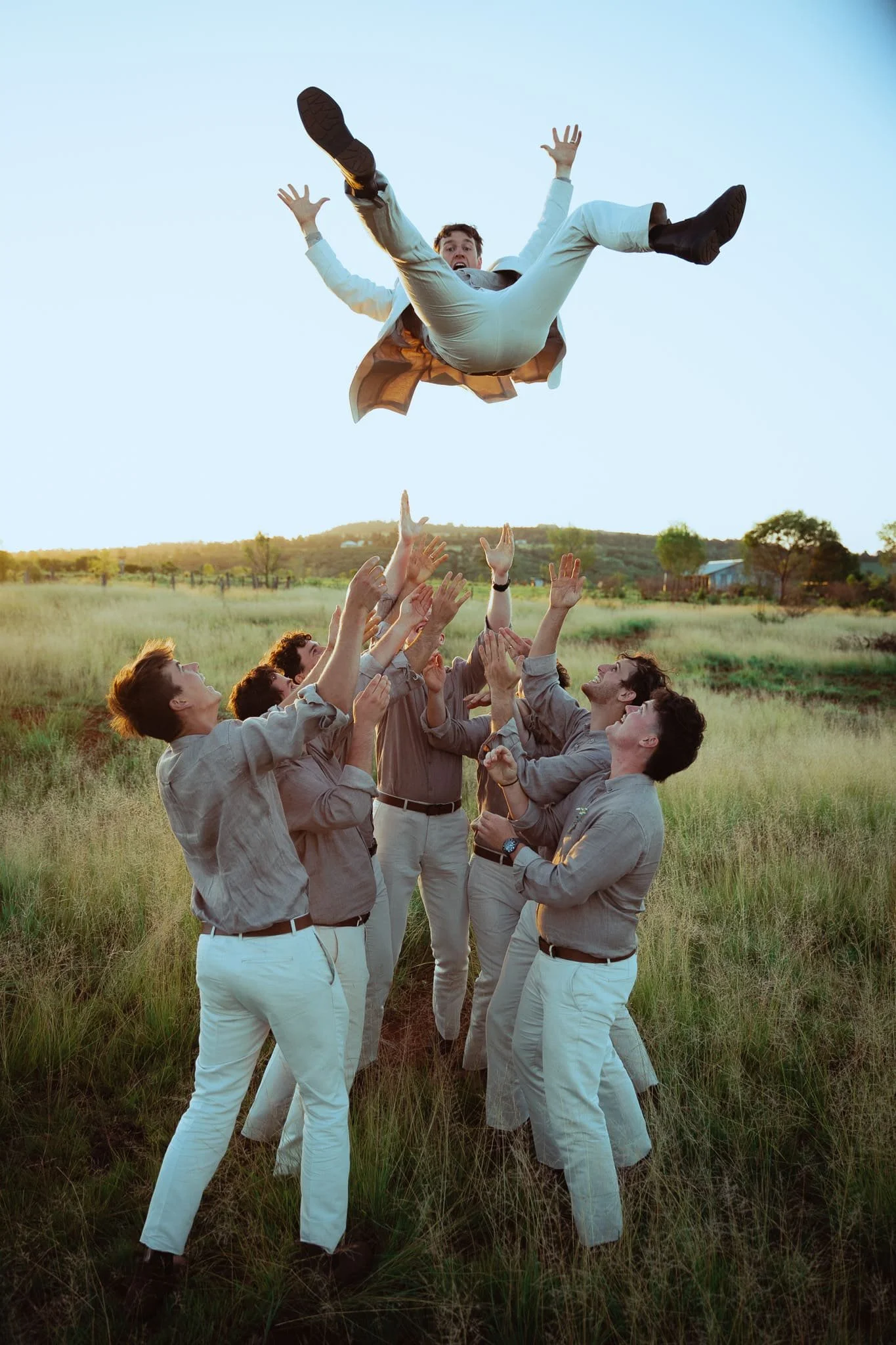 A man falling from the sky towards a group of people standing in a field at sunset, with their hands raised. The man has a surprised expression and is wearing a beige jacket, white shirt, and black boots.