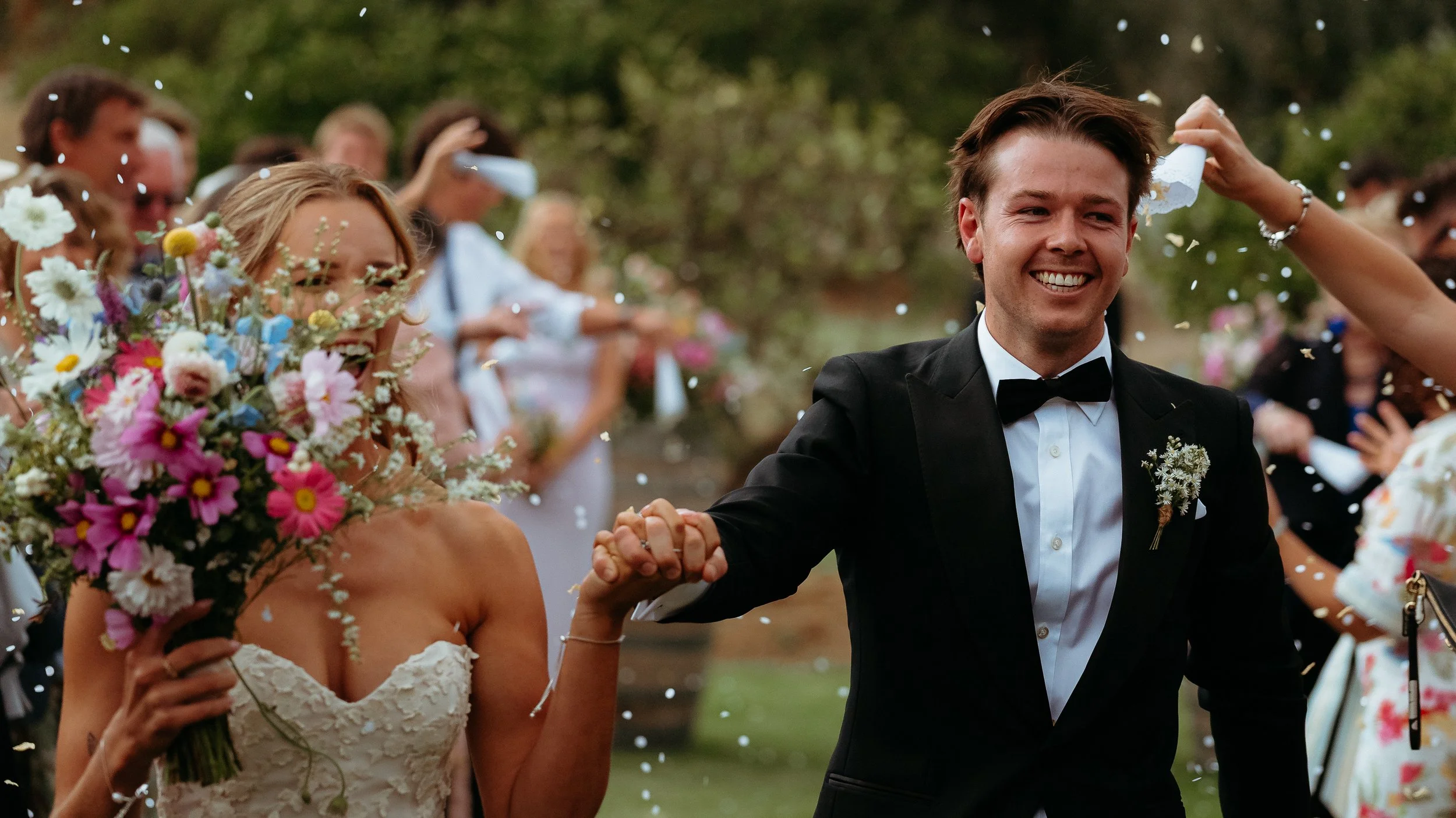 A smiling groom in a black tuxedo and bow tie holds hands with a bride in a strapless wedding dress as they walk through a celebration with guests and confetti outdoors.