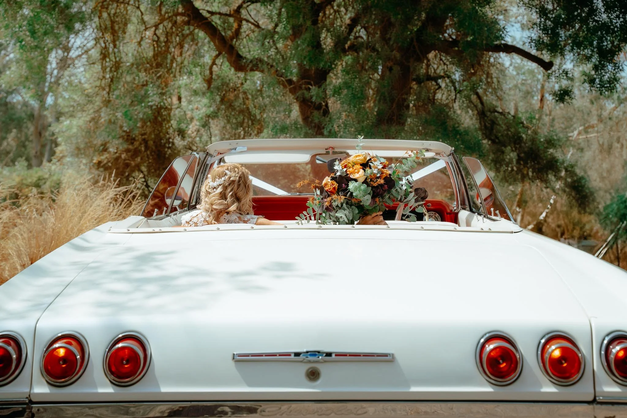 A bride sitting in a vintage white convertible car decorated with a large bouquet of flowers on the dashboard, surrounded by nature with trees and grass in the background.