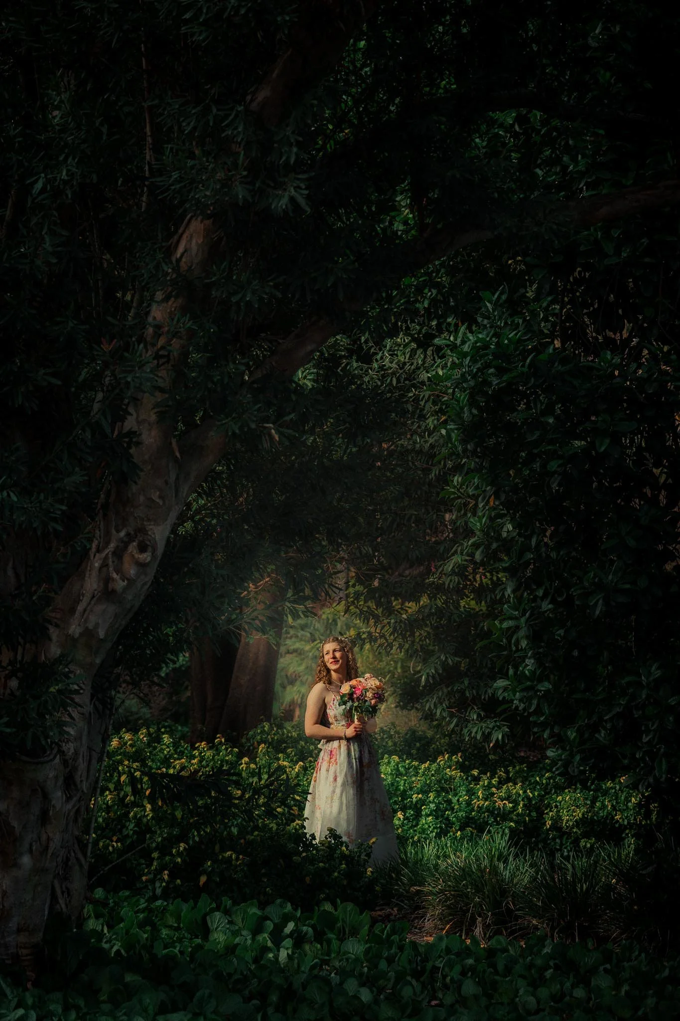 A woman in a floral dress holding a bouquet of flowers standing in a lush, green forest.