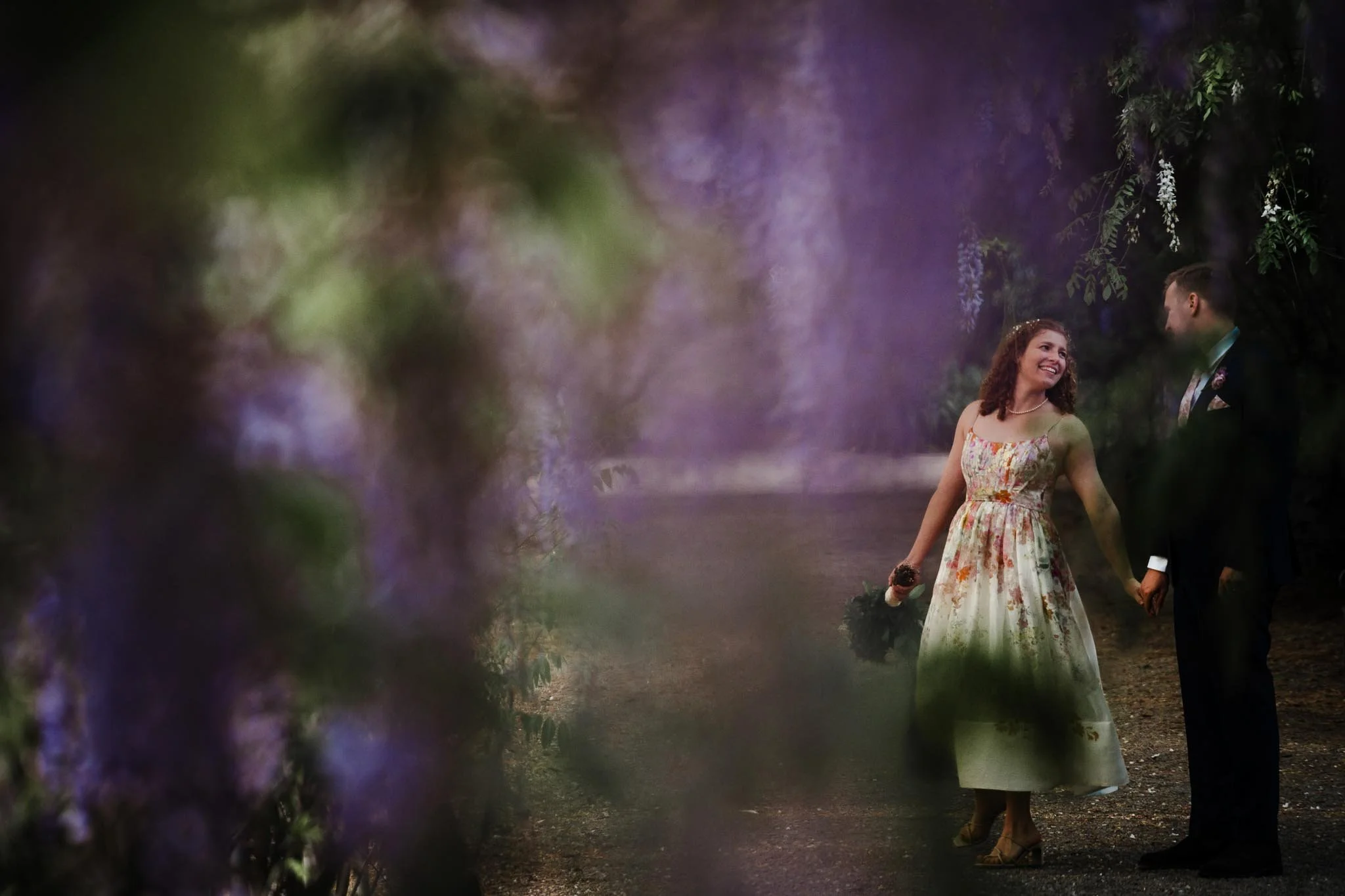 A couple in wedding attire holding hands and smiling at each other, walking on a path surrounded by lush greenery and purple flowers.