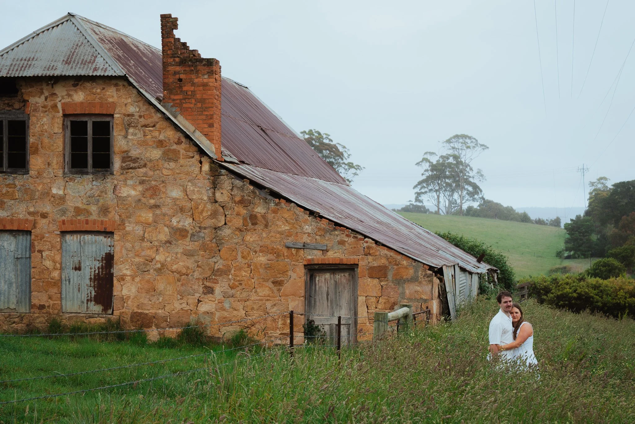 A rustic stone house with a rusted metal roof and boarded-up windows, set in a rural landscape with trees and a grassy field. Two people are standing outside the house, embracing.
