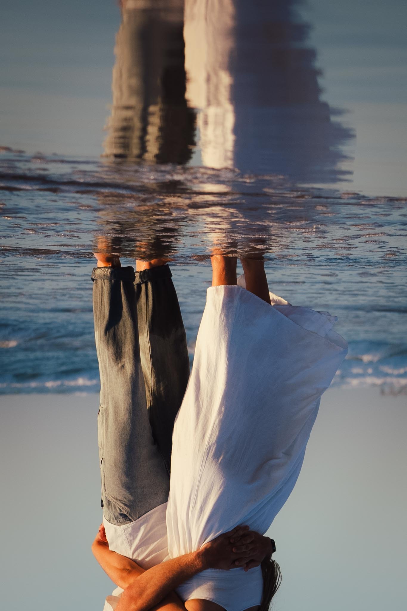 A woman wearing a white dress standing on the beach, with her reflection visible on the wet sand. The photo is taken during sunset or sunrise.