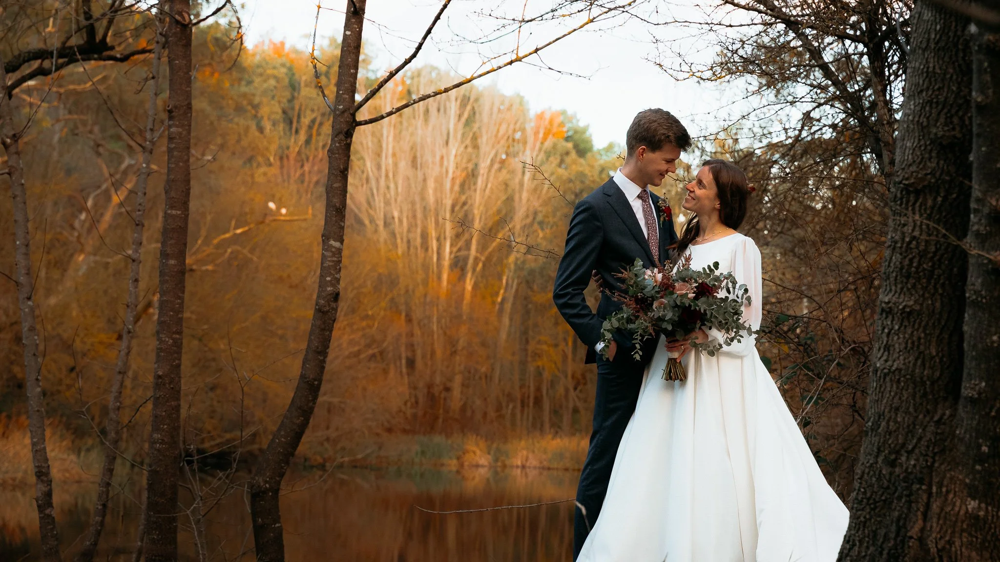 A bride and groom stand closely together outdoors during autumn, surrounded by trees with orange and yellow leaves. The bride wears a white wedding dress and holds a large bouquet of flowers, while the groom is dressed in a dark suit with a tie. They look at each other affectionately near a body of water.