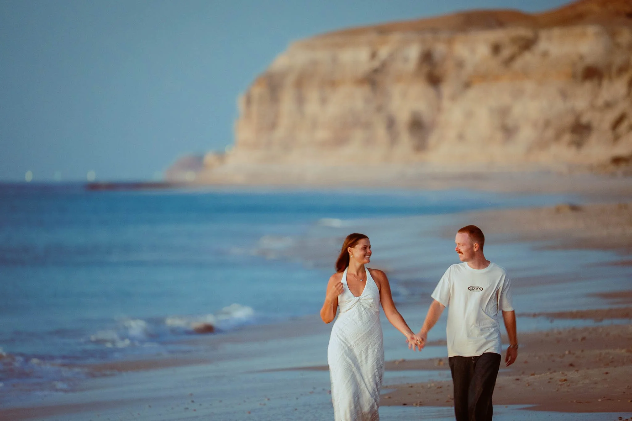 A couple holding hands and walking along the beach at sunset, with a large cliff in the background.