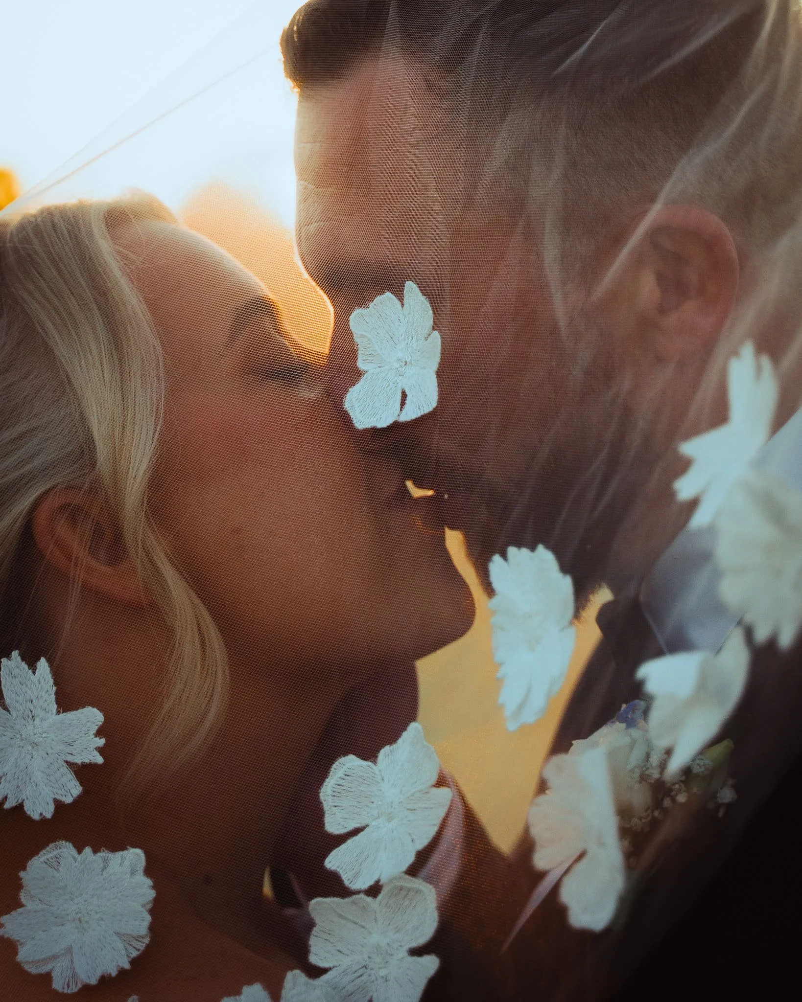 Close-up of a woman's face with floral lace veil, with the sun setting behind her, creating a warm glow.