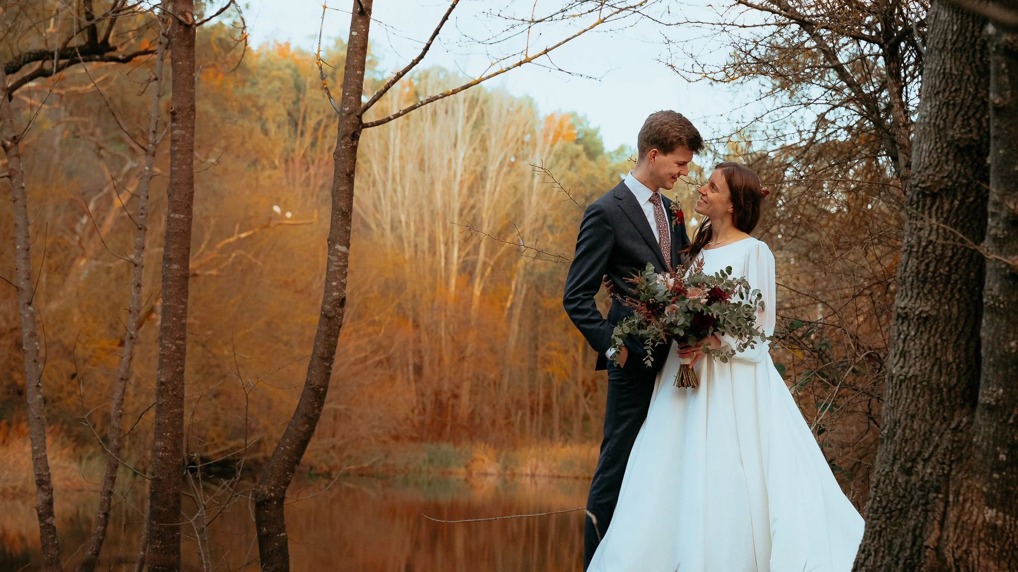 A bride and groom stand close together in a wooded area during autumn, with trees displaying fall foliage in the background. The bride is holding a bouquet of flowers, and they are smiling at each other.