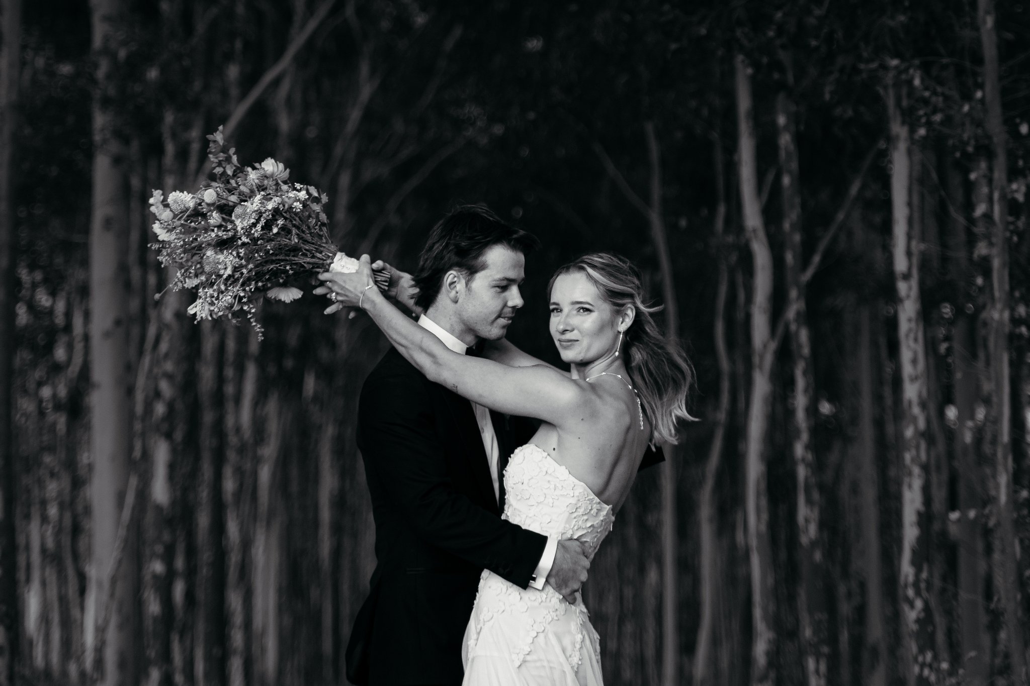 A black-and-white photo of a couple in wedding attire, with the man holding a bouquet of flowers behind the woman's shoulders, standing in front of a background of trees.