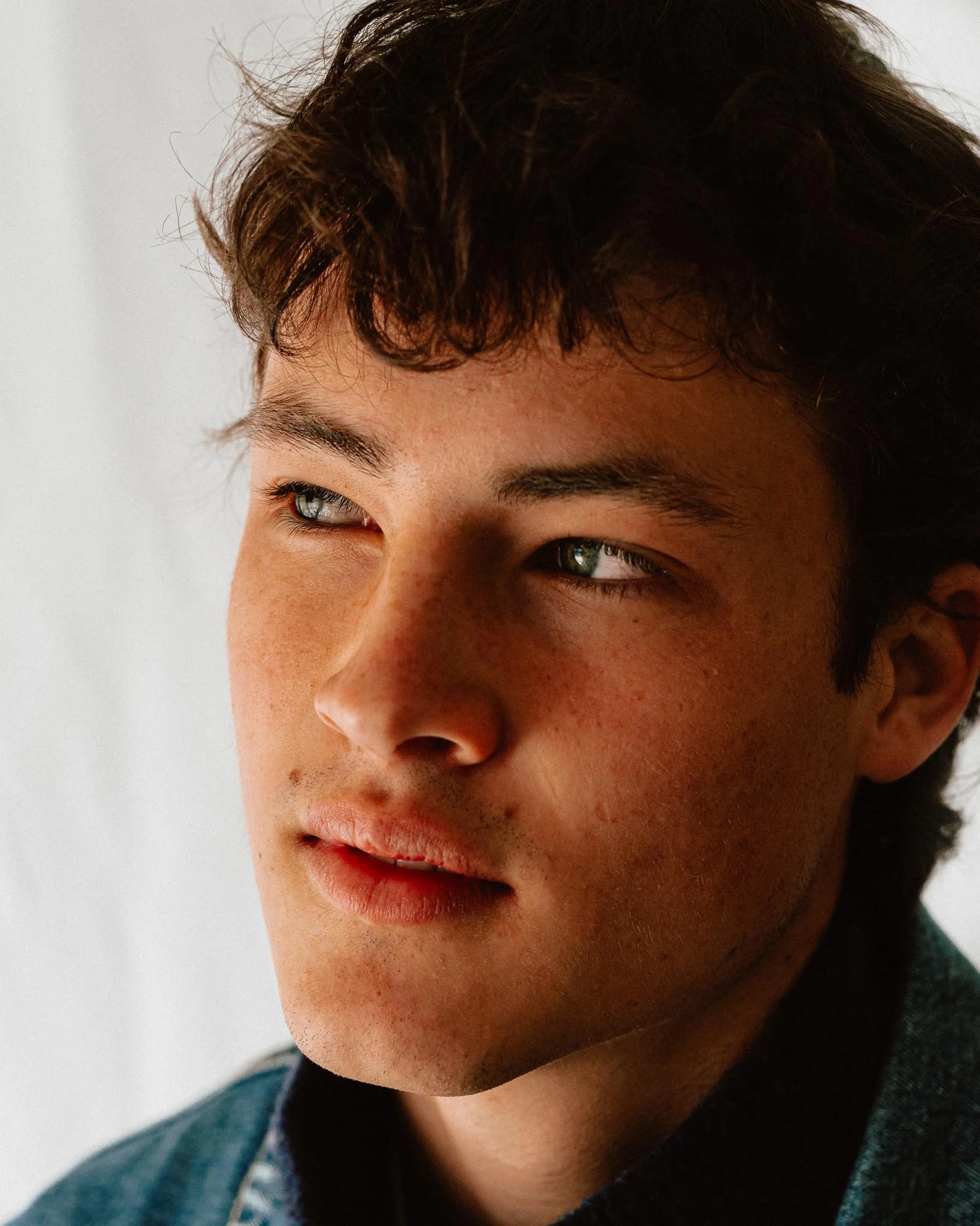 Close-up of a young man with dark curly hair, light skin, freckles, and green eyes, looking slightly to the side.