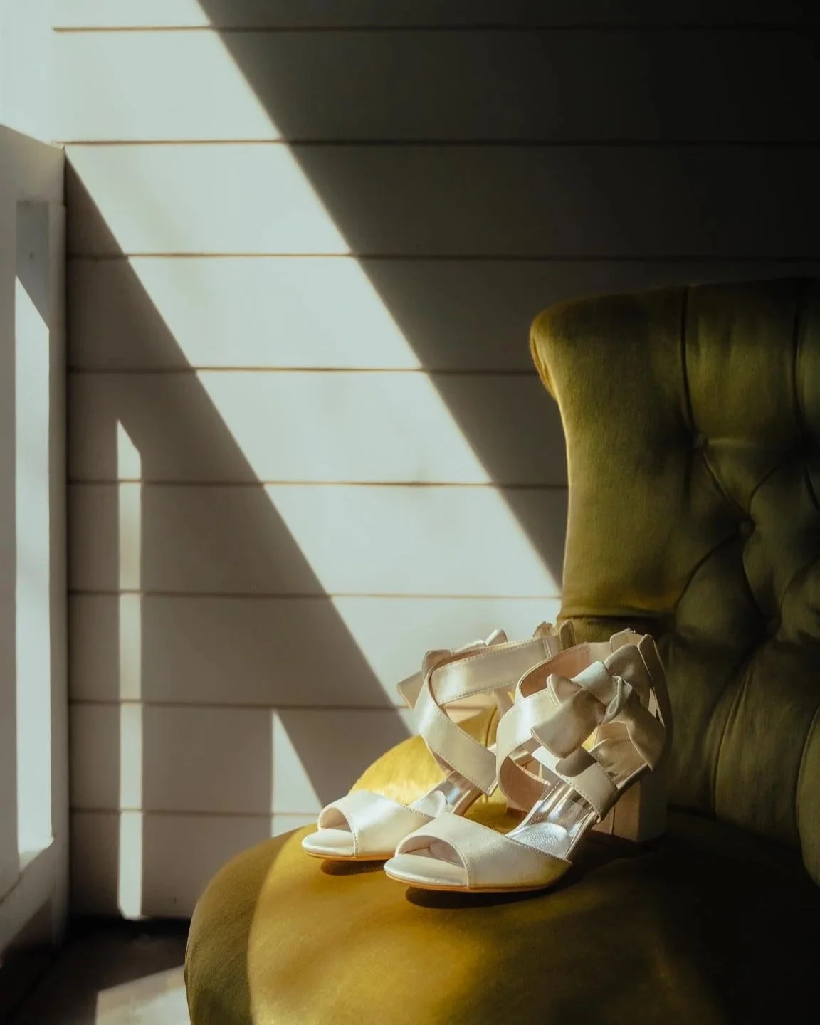 A pair of white high-heeled sandals with bows, placed on an olive green upholstered chair near a window with sunlight casting shadows.