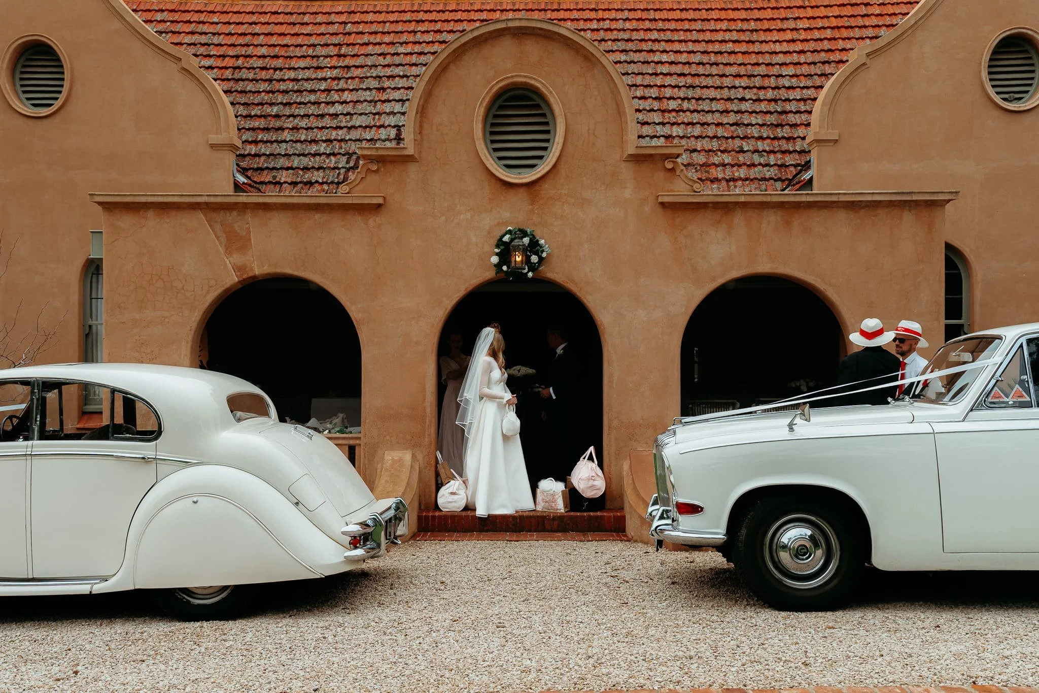 A wedding scene outside a tan stucco building with a red tiled roof. There are vintage white cars parked in front. A bride and groom are standing under the archway, with guests nearby, dressed in formal attire including hats and sunglasses.
