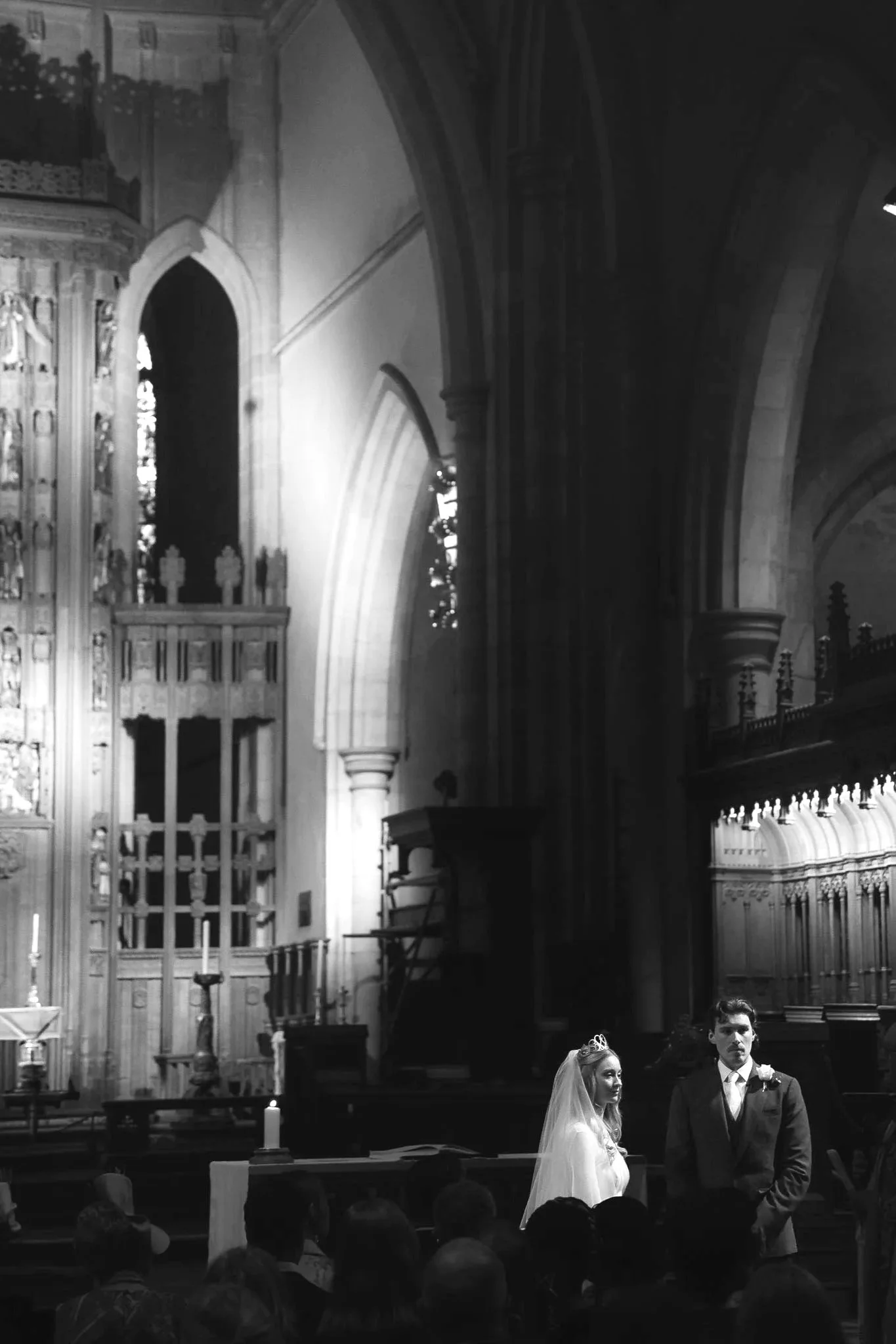 A black and white photo of a wedding ceremony inside a church. A bride and groom stand facing each other, with the bride wearing a white wedding dress and a tiara, and the groom in a dark suit with a boutonniere. The altar is decorated with candles and intricate woodwork, with seated guests visible in the foreground.